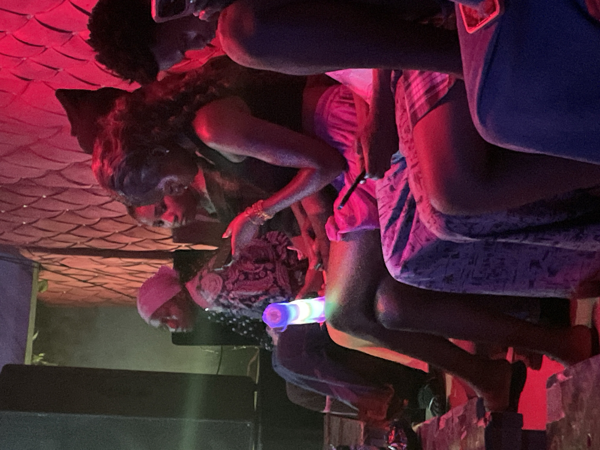 In Durumi, Nigeria, a group of individuals are seated in a dimly lit indoor setting, possibly a lounge or club, characterized by red ambient lighting and textured walls. In the foreground, a person in the lower right is partially visible, seated and dressed in blue. To their left, another individual is seated, with their legs crossed, wearing a pink and white plaid skirt and a dark top. Their right hand holds a black smartphone.
Further back, a person with long, curly dark hair is seated, wearing a dark top and a bright pink skirt. Their left arm is extended, resting on their leg, and they are holding a small black object, possibly a phone. To their left, another person is visible with their head turned away, wearing a patterned fabric, possibly a headscarf, and a patterned garment. Behind them, a person in a pink headscarf is seated, looking to the left.
In the background, to the left, a large dark speaker or structure is present. A glowing, multi-colored object, resembling a light-up cylinder, stands upright on a surface between the people. The overall atmosphere appears to be social, with a focus on seated individuals interacting within a club-like environment.