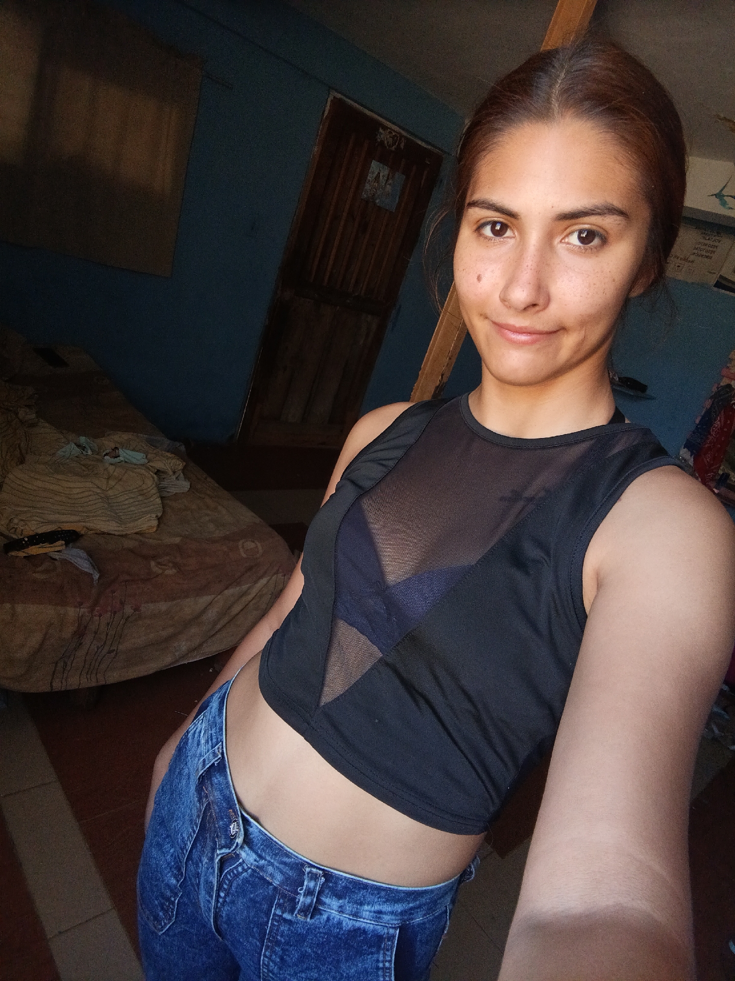A young woman is shown from the chest up, taking a selfie in what appears to be a bedroom. She has light brown hair pulled back, fair skin with freckles, and is wearing a black mesh tank top over a dark bra and blue jeans. She is smiling and looking at the camera. The background is dimly lit, with a blue wall, a wooden door, and a bed with a rumpled beige blanket visible to the left. The room appears to be in a modest dwelling.