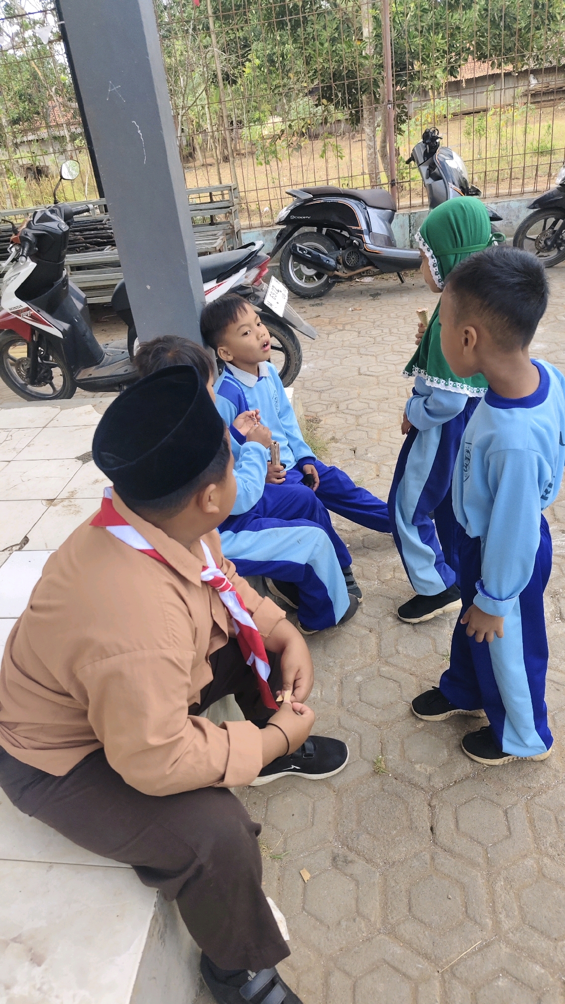 The image shows a group of children in light blue uniforms and a boy in a brown uniform with a red and white scarf, seated and standing near motorcycles outside. The scene appears to be in a public area, possibly a schoolyard or community space, with a paved ground and a metal fence in the background. The children are engaged in conversation, with some looking at each other and one child holding a snack. The motorcycles, a Honda and a Yamaha, are parked nearby. The environment is outdoor, with trees and foliage visible in the background. The lighting suggests it is daytime.