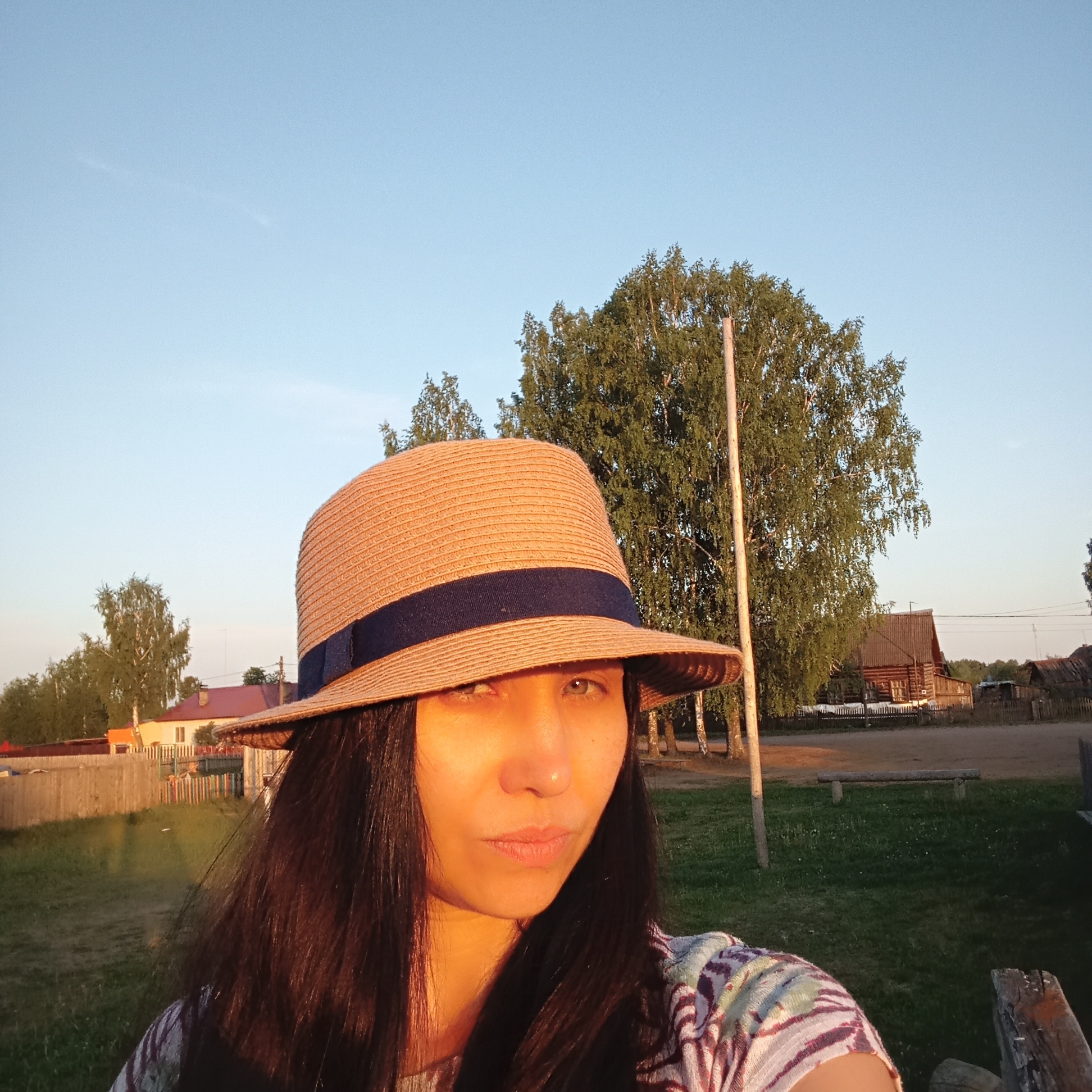 A woman wearing a brown fedora with a dark blue band is centered in the image. She has long, dark hair and a patterned shirt. The background shows a rural setting with houses, trees, and a blue sky. Based on the information given, the location is the city of Буренка, Russia. A wooden bench is visible in the background, along with a fence and a dirt road. The lighting suggests it is either sunrise or sunset.