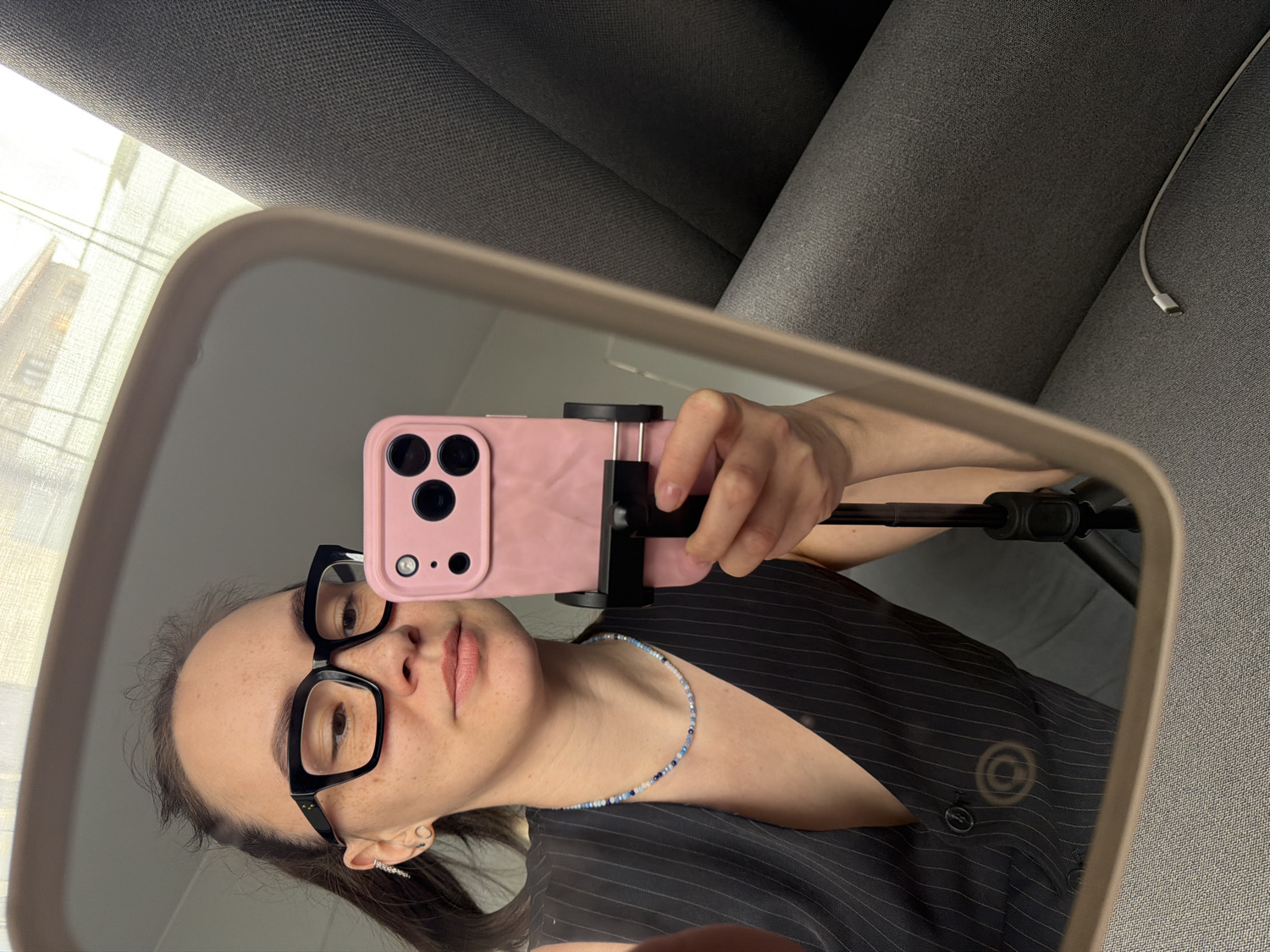 A mirror selfie captures a woman's reflection, showing her holding a pink-cased smartphone mounted on a black tripod or selfie stick. She wears black-framed glasses, a dark pinstriped vest or top, and a light blue beaded necklace. Her dark hair frames her face, which has visible freckles, and she looks directly into the camera with a neutral expression.
The indoor setting is reflected in the mirror. Behind the woman, a grey upholstered sofa is visible, with a white charging cable resting on it. A bright window or light source, partially obscured by a light curtain, can be seen in the upper left corner of the reflection, indicating daytime. The activity is the woman taking a self-portrait using a mirror and phone stand. The scene appears to be set in a domestic environment, such as a home in Chaikovsky, Russia. No discernible text is visible in the image.