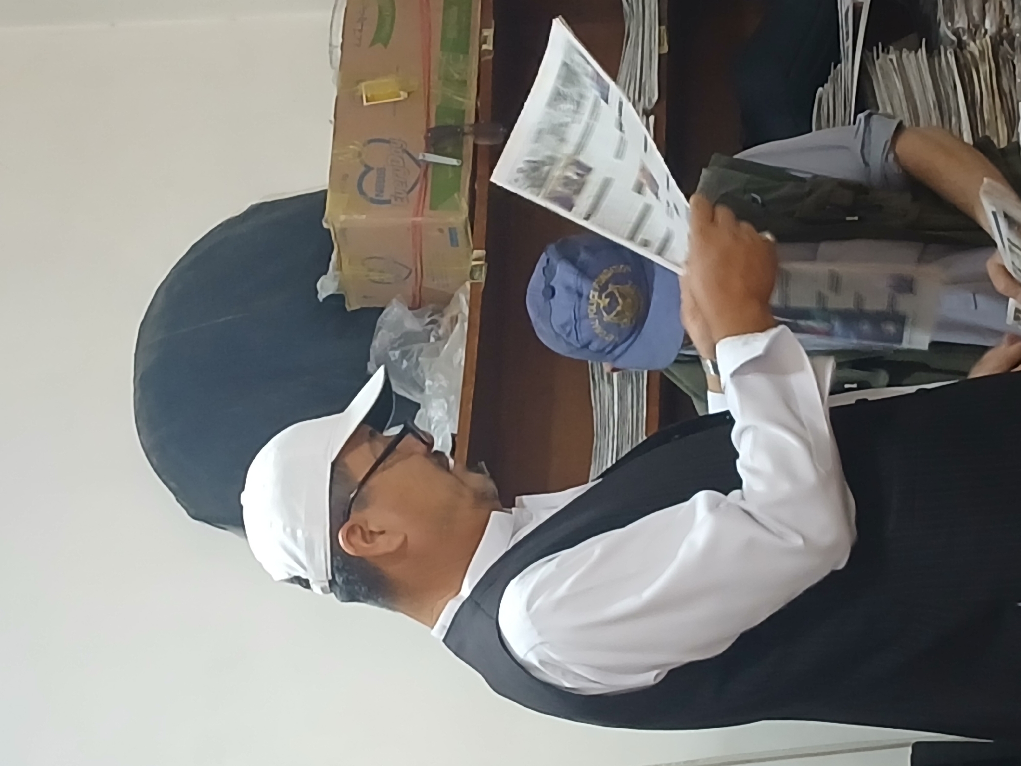 A man is reading a document indoors. The man has a dark complexion, a white cap, and glasses, and he is wearing a black vest over a white long-sleeved shirt. He appears to be reading a document with pictures in his hands. A blue baseball cap with a yellow crest is visible, sitting next to the man's arm. Behind the man is a brown shelf with what appears to be several files or documents stacked in it, along with a box of what appears to be Nestle Milo, a brand of beverage. There is also a black object leaning against the shelf. The location is Quetta City Tehsil, Pakistan.
