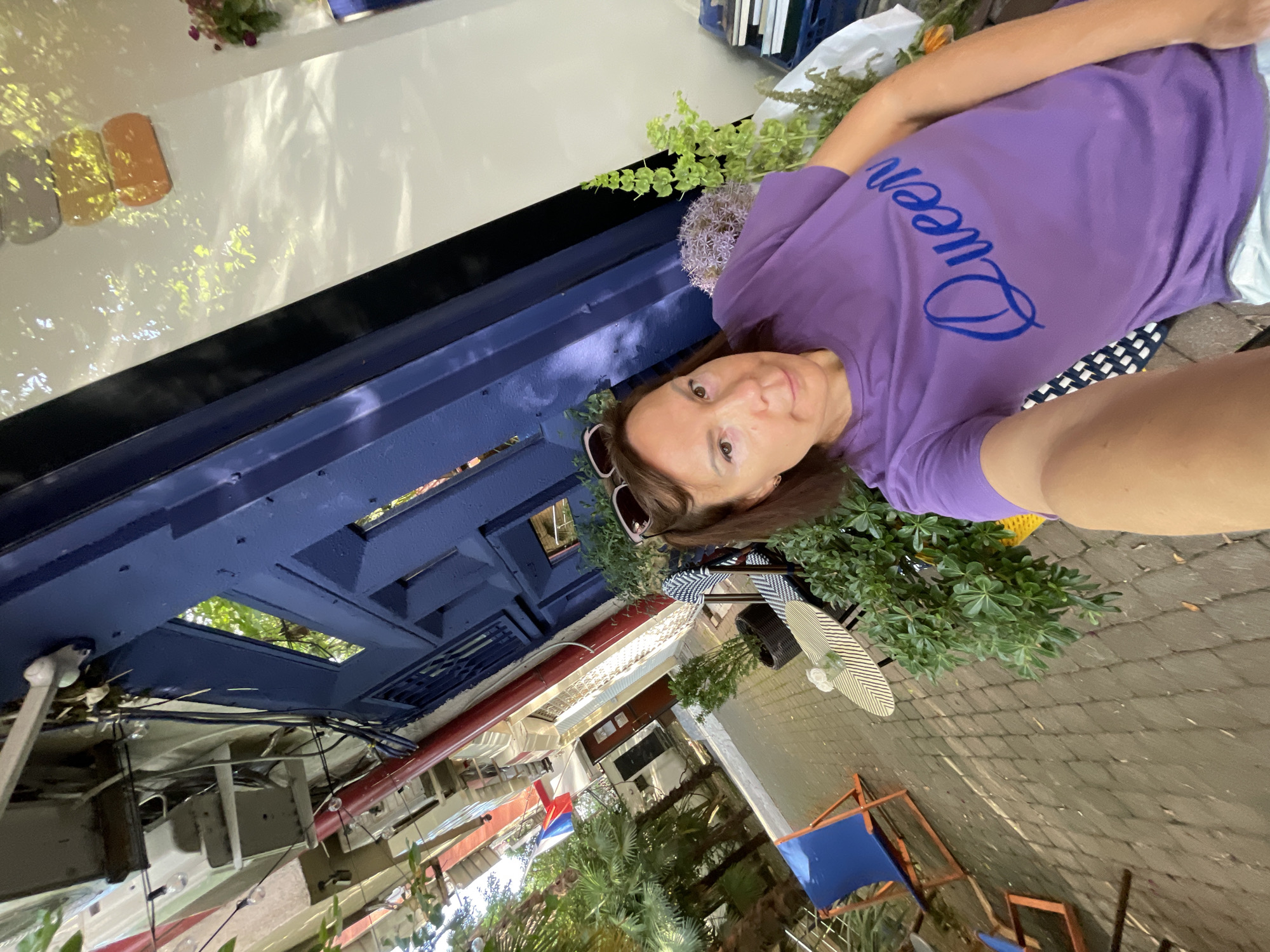 A woman is taking a selfie outdoors in front of a building with blue painted architectural elements. She is wearing a purple t-shirt with "Queen" written on it in blue. Sunglasses are perched on top of her head. She is seated on a chair with a black and white patterned seat. Behind her, there are plants, a table with a glass of water, and chairs. In the background, there are more plants, palm trees, and what appears to be part of a building with red and white accents. The ground is paved with cobblestones. The scene suggests a cafe or restaurant setting, likely in a warm climate.