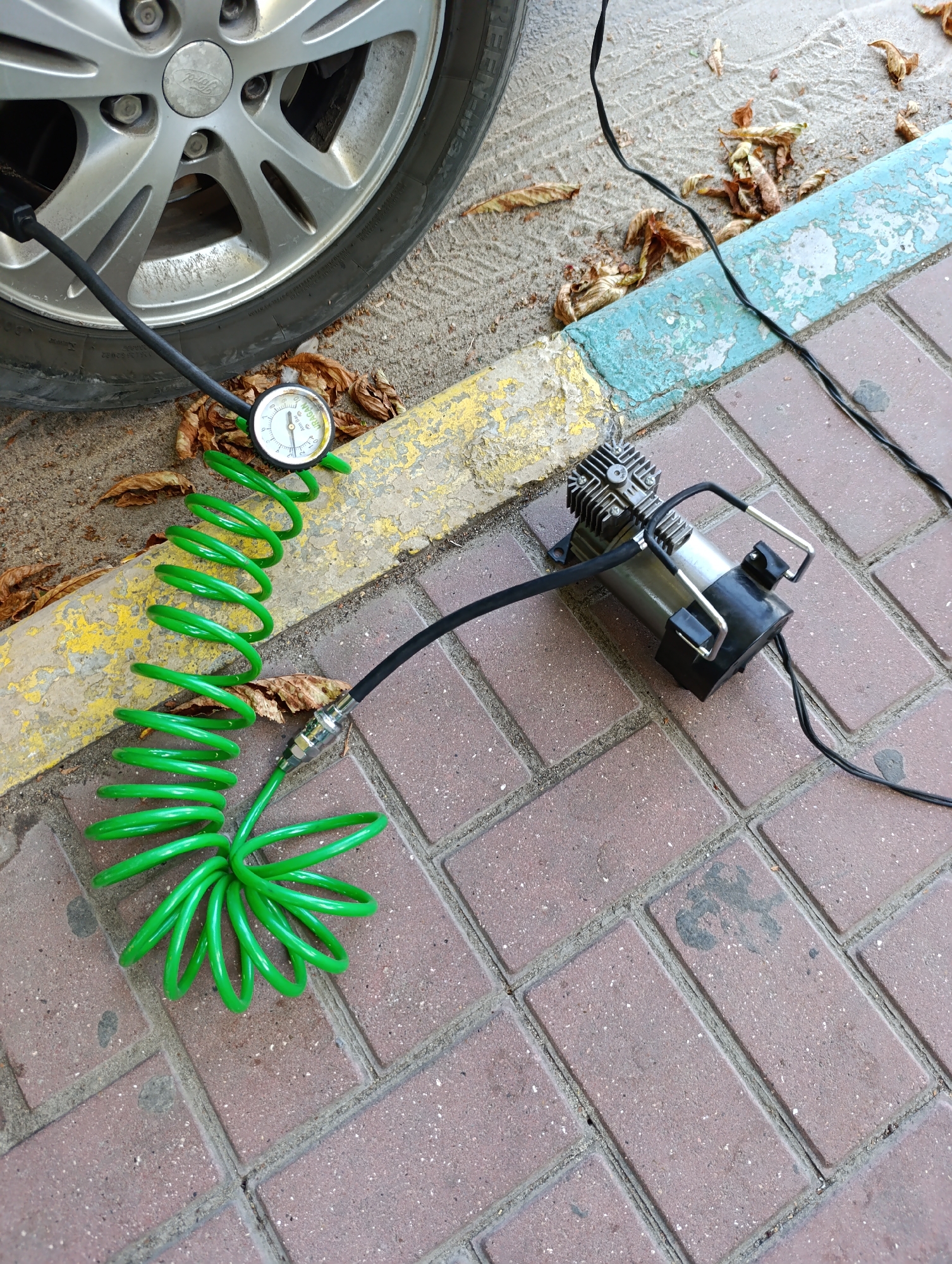 An outdoor, eye-level, medium shot shows a car tire and a portable tire inflator on a brick-paved surface. The car tire is on the left, with its silver rim visible and the tire pressure gauge attached to the valve stem. A bright green, coiled hose connects the gauge to a compact, silver and black tire inflator, which is situated to the right of the tire. The inflator is plugged into a black power cord that runs across the pavement. The scene is set on a sidewalk or road, with a curb painted yellow and blue in the background. Fallen brown leaves are scattered on the ground, suggesting an outdoor setting. The image is a still from a video feed, capturing a moment of car maintenance. The location is the town council of Dubăsari, Moldova.