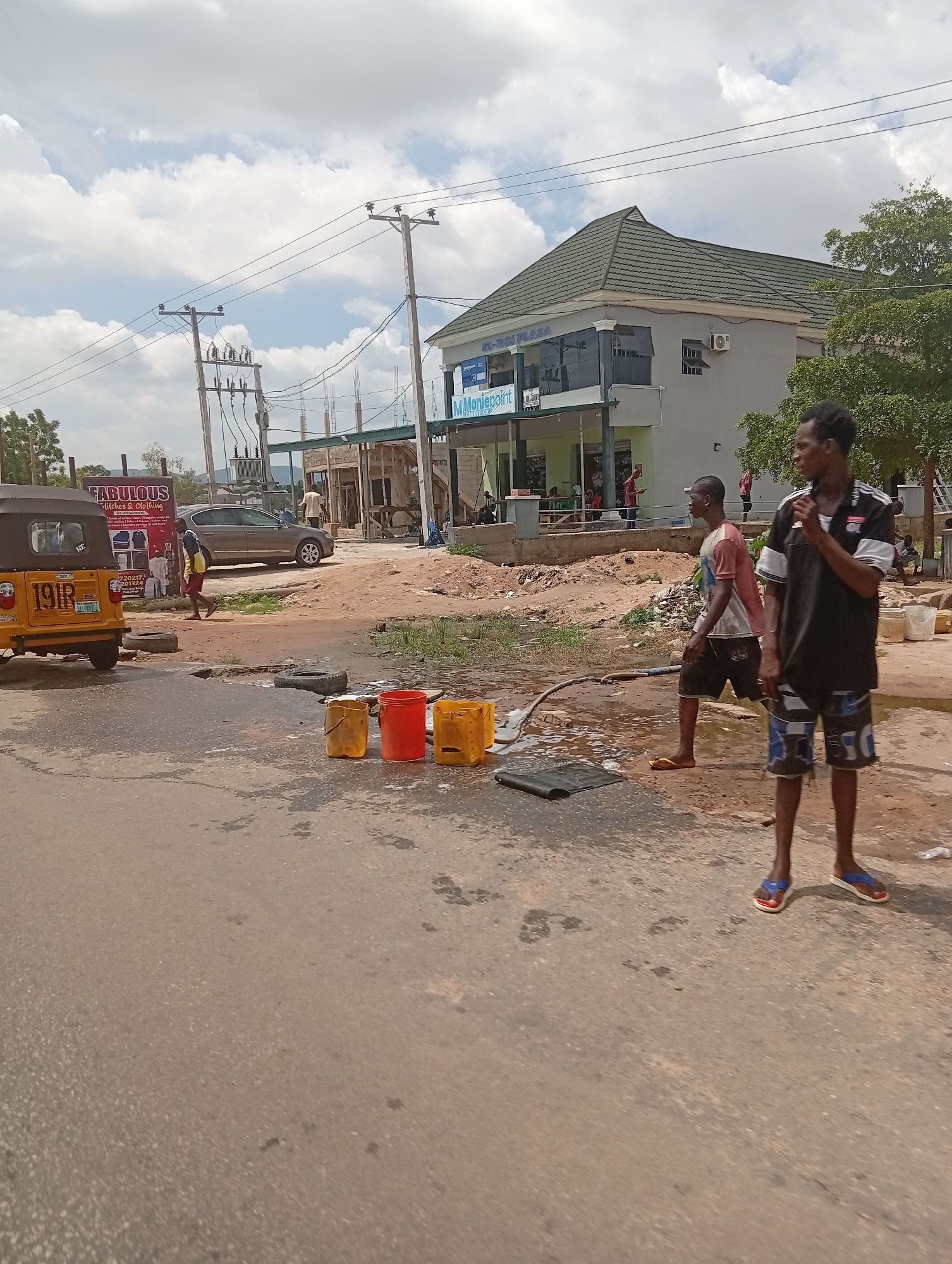The image captures a scene on a street in Jalingo, Nigeria, likely during daylight hours given the bright sky and shadows. In the foreground, the paved road is wet, with several yellow and red plastic containers, possibly for collecting water, placed on the wet surface. A black mat lies near the containers. Two young men are visible. One, on the right, is wearing a black and white t-shirt and patterned shorts, and appears to be holding something to his mouth. The other, to his left, wears a red and white t-shirt and dark shorts, and is walking. A hose is visible connecting to the ground where water is pooling.
In the mid-ground, a yellow motorized tricycle, or 'Keke NAPEP', with the number plate "19R" is parked on the left. Behind it, a gray car is parked. A billboard for "FABULOUS Stitches & Clothing" is also visible. Further back, a building identified as "EL-ROI PLAZA" is prominent, with "MMoniepoint" signage. Construction appears to be ongoing around this building, with visible scaffolding and building materials. Overhead, utility poles with electrical wires crisscross the sky. The environment is dusty and somewhat unkempt, with scattered debris. Trees line the background.