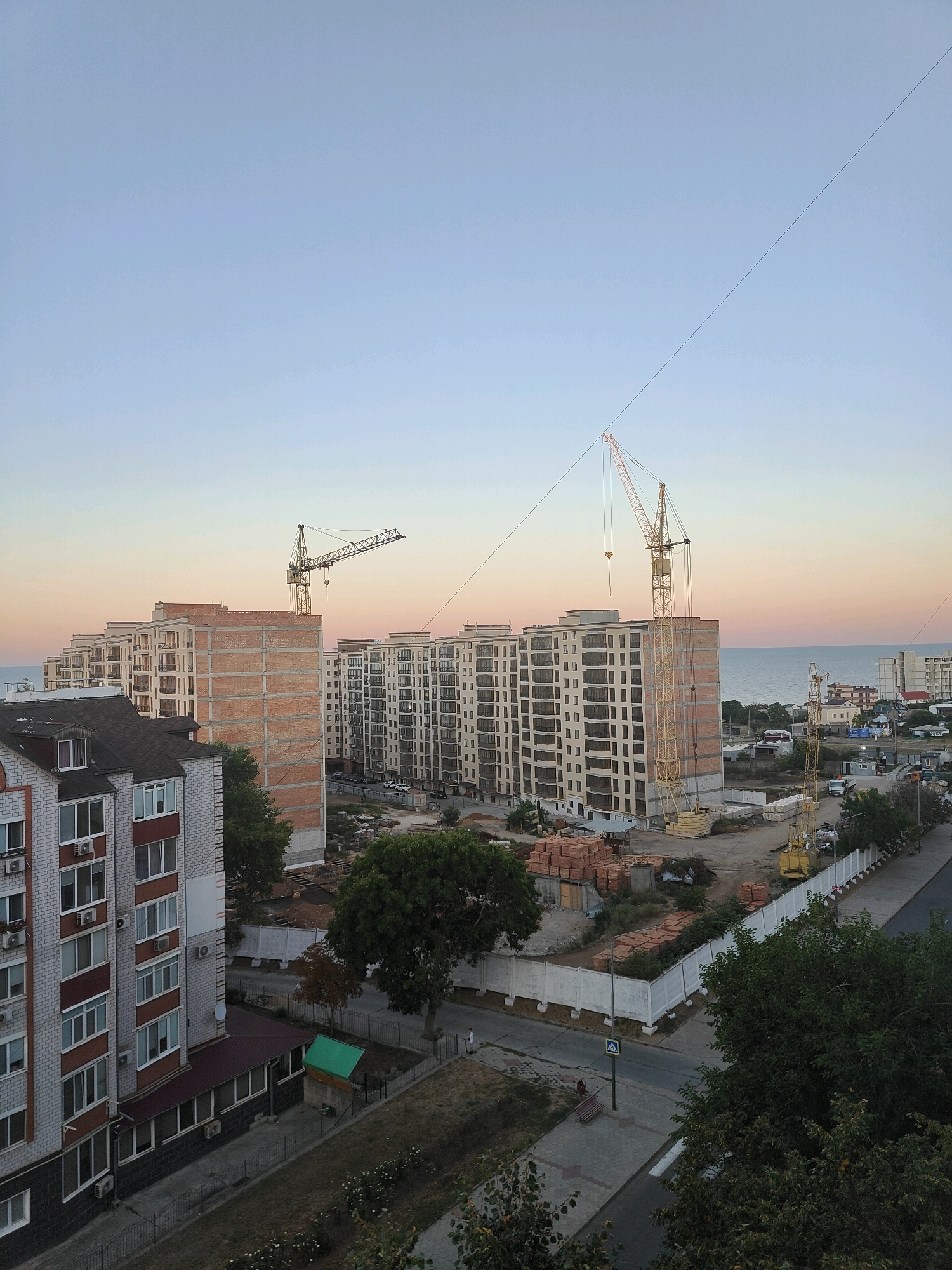 This is an aerial, eye-level, medium shot of a construction site in Chornomorsk, Ukraine, at sunset. The sky is a gradient of pale blue to pink. In the foreground, a multi-story residential building with white and brick facades and visible air conditioning units is partially visible on the left. Adjacent to it, a larger, multi-story building under construction, primarily brick, is visible. Two large yellow tower cranes are present on the construction site, with one extending over the partially built structure. Several stacks of bricks and construction materials are visible on the ground. Beyond the construction site, more residential buildings and a road with a few vehicles are visible. In the distance, the Black Sea meets the horizon. A lone pedestrian is visible on the sidewalk in the lower right portion of the image. The overall scene depicts ongoing urban development.