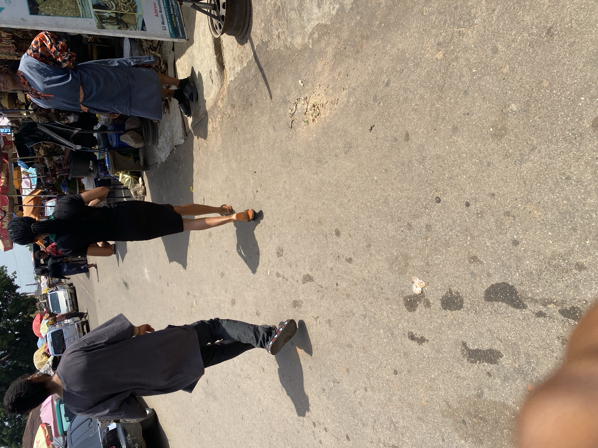 The image depicts a street scene in Jos, Nigeria, likely a marketplace or a busy thoroughfare. In the foreground, three individuals are visible from behind, walking away from the camera. The person in the center, wearing a black dress, is most clearly defined, with their back to the viewer, displaying a braid styled ponytail. To their left, a person in a dark grey t-shirt and dark pants is walking, their head turned slightly to the left. To the right of the person in black, another individual is seen from the waist down, wearing a blue and orange patterned garment over a dark skirt, with their right hand on their hip.
The environment is paved with asphalt, showing signs of wear and tear, with scattered debris, including what appears to be dried food scraps and possibly water stains. In the background, various stalls with colorful awnings are visible, suggesting a market setting. Several vehicles, including a white minivan and a darker vehicle, are parked or driving on the street. The overall impression is of a sunlit, active outdoor space in an urban area of Nigeria.