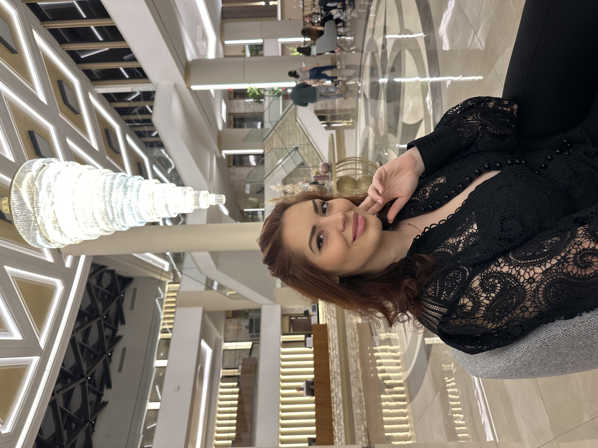 The image captures a young woman posing in a modern, brightly lit indoor space, likely a hotel lobby or a multi-level public building in Kazanlak, Bulgaria.
The primary subject is a young woman, positioned on the right side of the frame, looking directly at the viewer with a pleasant, soft smile. She has reddish-brown hair and is wearing a black long-sleeved top with intricate lace patterns and a deep V-neck, adorned with black buttons. Her right hand is gently placed near her jawline.
The background reveals an expansive and contemporary interior. A large, multi-tiered chandelier, made of numerous crystal or glass elements, hangs prominently on the left, casting bright white light. The ceiling features geometric panels with integrated linear lighting, creating a sophisticated design. The floor is polished, displaying a complex circular and geometric pattern.
In the mid-ground and background, several other people are visible, some seated in lounge areas and others appearing to walk through the space, indicating a public and potentially busy environment. Architectural elements include large, light-colored pillars, glass railings for upper levels, and stairs connecting different floors. The overall setting suggests a modern and upscale design. No visible text is discernible within the image.