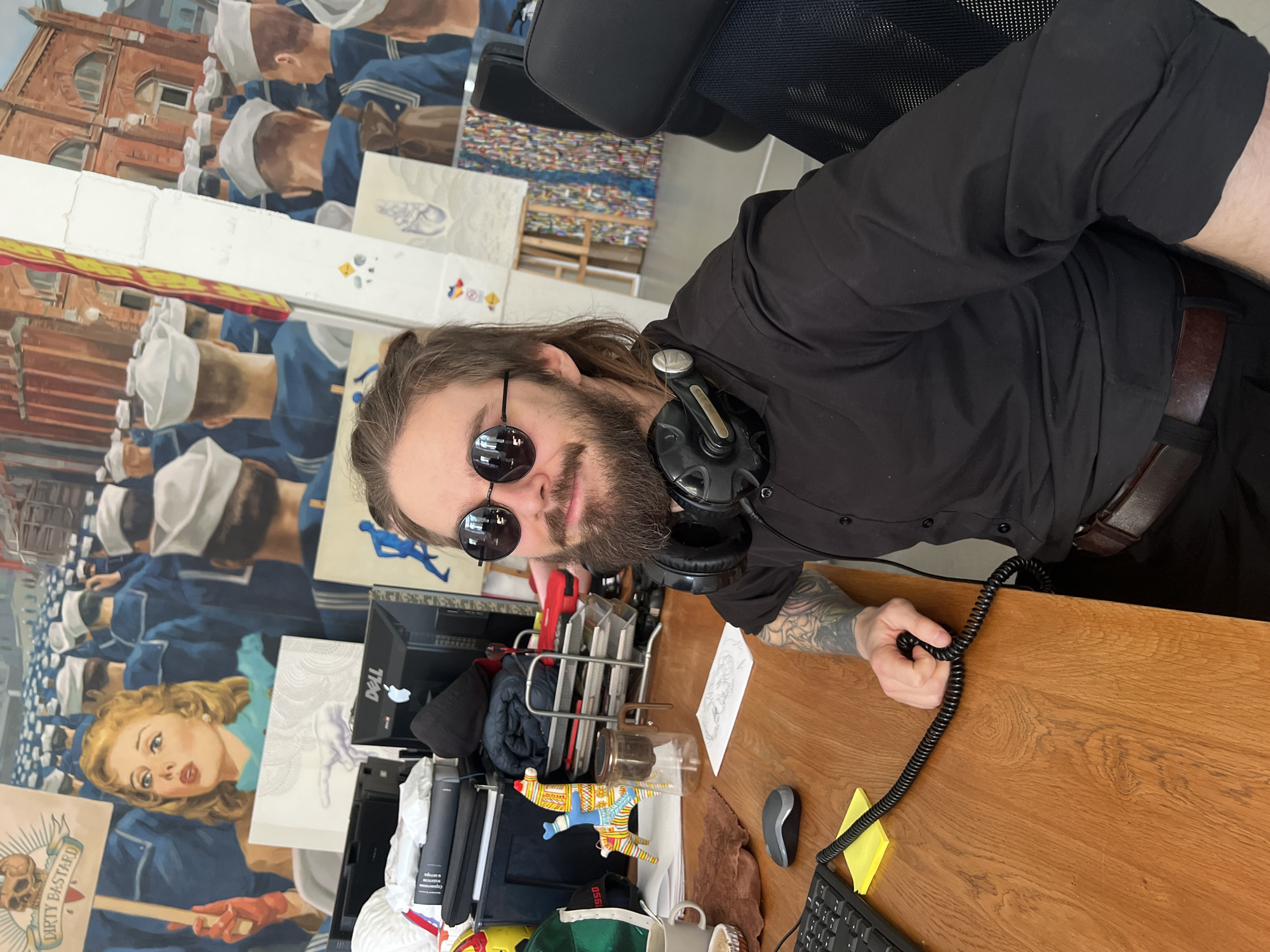 The media file captures a man with long brown hair and a beard, wearing round sunglasses and a black collared shirt, looking directly at the camera with a slight smile. He has a pair of black headphones resting around his neck and visible tattoos on his left forearm. He is seated at a wooden desk, holding a coiled black cord in his right hand.
The background is dominated by a large, colorful mural. It depicts several men in white sailor hats and blue uniforms, seemingly marching or queuing, with parts of a red brick building visible above them. In the lower left of the mural, a blonde woman with red lips and red gloves holds a sign that reads "DIRTY BASTARD" and features a skull and crossbones emblem.
The desk holds various items, including a computer mouse, a keyboard, yellow sticky notes, a clear glass pour-over coffee maker, a colorful llama-shaped decoration, a white mug, and a black baseball cap with "1950" partially visible. A "Dell" logo and an Apple logo are visible on objects behind the desk organizer. Further in the background, stacks of artworks or canvases can be seen. The setting appears to be an office or art studio, suggesting a creative environment in Saint Petersburg, Russia. The man's expression is relaxed, and the scene is brightly lit, indicative of daytime.