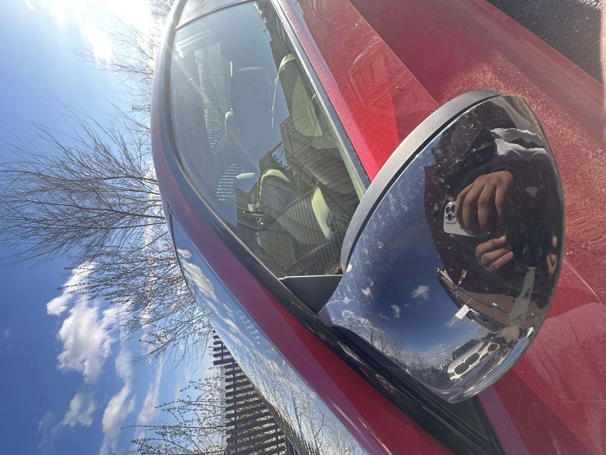 A close-up, canted shot focuses on the side mirror of a red car, reflecting a person holding a phone, a building, and a white vehicle. The car is dusty. The reflection in the mirror also includes blue sky and bare branches. The foreground shows the red body of the car. The environment suggests an outdoor setting, possibly a street or parking lot.