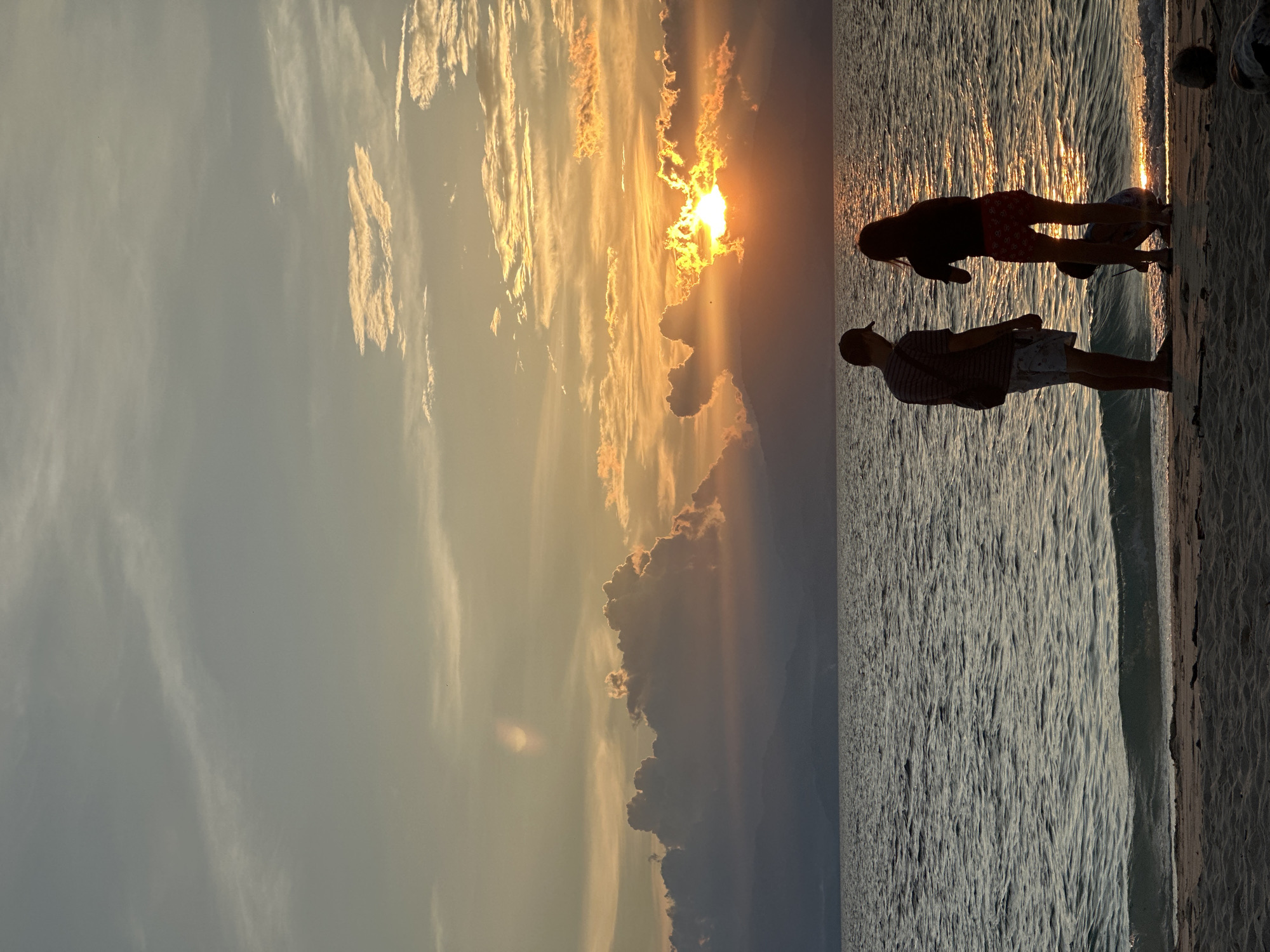 A group of people are silhouetted on a sandy beach facing a body of water during sunset. The sky is filled with colorful clouds, with the sun partially obscured behind them, casting a warm glow. The water is calm with gentle waves lapping at the shore. The people appear to be enjoying the view, with two standing and one crouching on the sand. The scene captures a peaceful moment at the beach, likely in Saavedra, Philippines, given the tropical setting and common tourist activities.