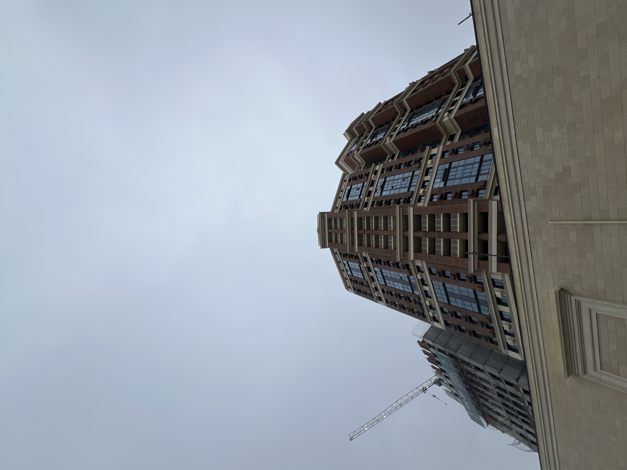The image presents an upward-looking view of several buildings under an overcast sky, captured with a rotated perspective.
In the foreground, dominating the left and central parts of the frame, is a tall, multi-story building with a brown facade. This building features a distinctive design with receding upper floors and a stepped or zig-zag pattern formed by balconies or recessed sections. Numerous rectangular windows, dark and reflective of the pale sky, are arranged in vertical columns. White or light-colored trim details adorn the edges of these sections and windows.
To the right of this prominent brown building, a large section of a lighter, beige or tan-colored building is visible. This structure has a textured, block-like facade, possibly made of brick or stone tiles, and appears to be an adjacent or nearby building with a simpler, more uniform design. Its top edge is not visible, and a decorative, framed relief can be seen on its lower right portion.
Further down and to the left of the main brown building, another building is visible, appearing to be under construction. This structure is grey, with visible scaffolding or protective netting, and a tower crane stands next to it, indicating ongoing building activity.
The sky is uniformly light grey and overcast, suggesting a cloudy day without direct sunlight. No people or clear textual elements are visible in the frame. The overall scene depicts a modern urban environment with a mix of completed and under-construction architecture.