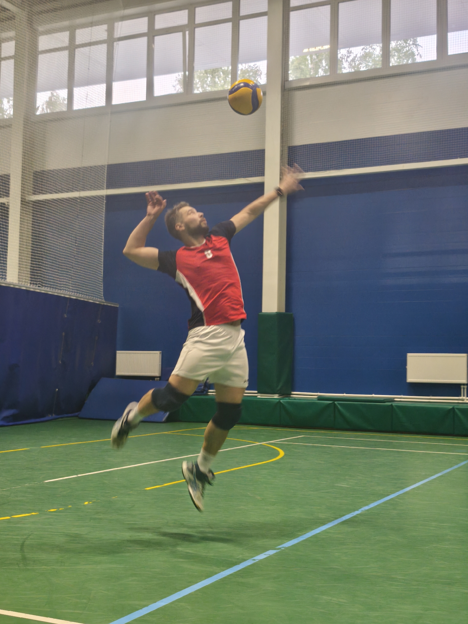 A man is jumping to hit a volleyball in an indoor sports arena, likely for a game or practice. He is wearing a red, white, and black sports jersey, white shorts, knee pads, and athletic shoes. He has a beard and his arm is raised to strike the yellow and blue volleyball. The arena has a green floor with yellow and blue lines indicating court boundaries, a blue wall, white support pillars, and windows near the ceiling. Based on the architecture, this could be a sports facility in Saint Petersburg, Russia.
