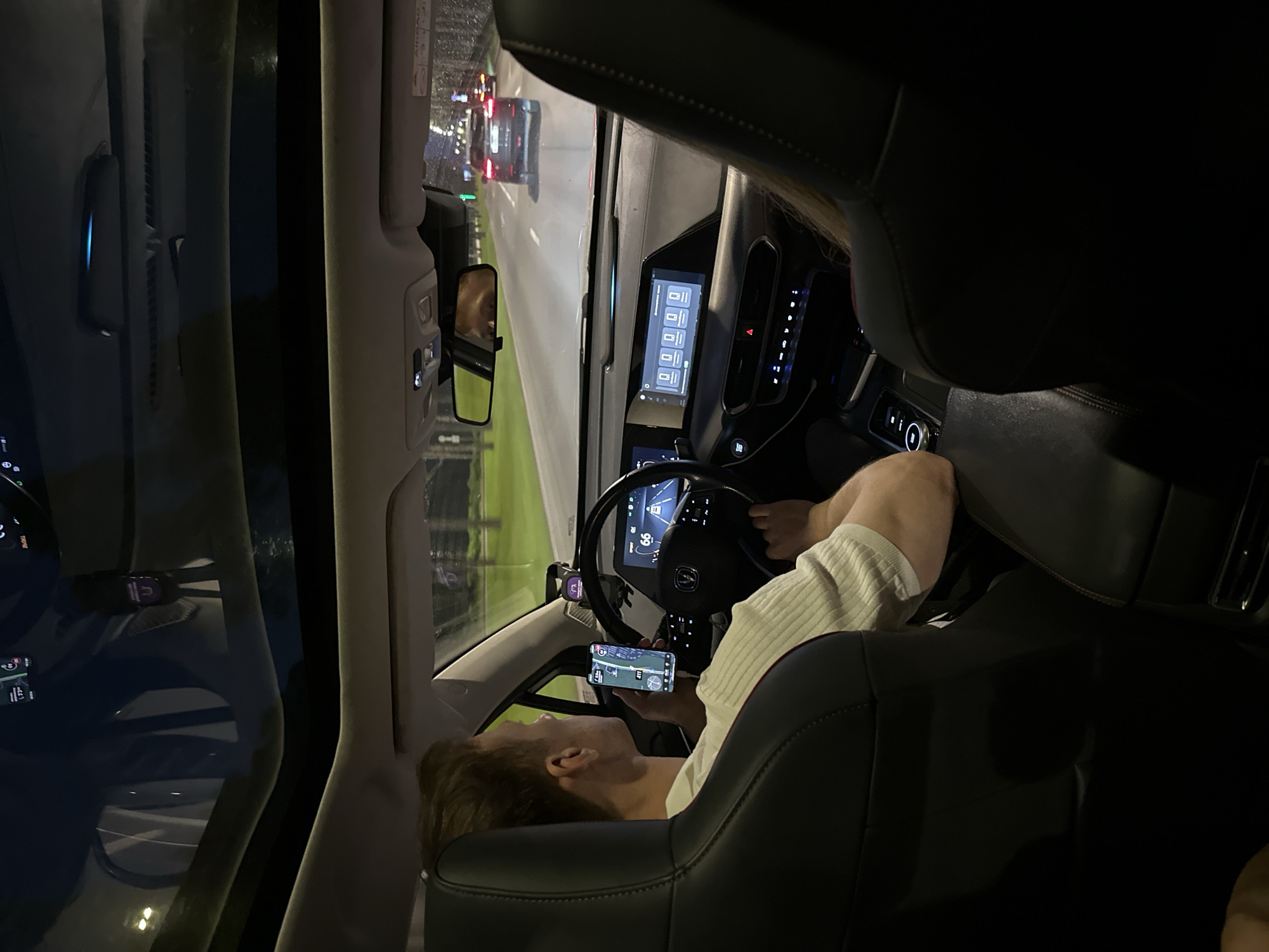 The image shows the interior of a car at night, looking forward through the windshield and sunroof. A man is driving, holding a smartphone displaying a navigation app. The car's dashboard features multiple screens, including one displaying speed and navigation. Another screen shows icons that appear to be related to media or settings. Outside the car, streetlights and trees are visible, indicating a suburban or urban environment. The road ahead shows other vehicles, suggesting the car is in motion. The overall lighting suggests it is nighttime, and the presence of rain is indicated by the streaks on the windshield. There is no explicit information to pinpoint the location to Saint Petersburg, Russia.