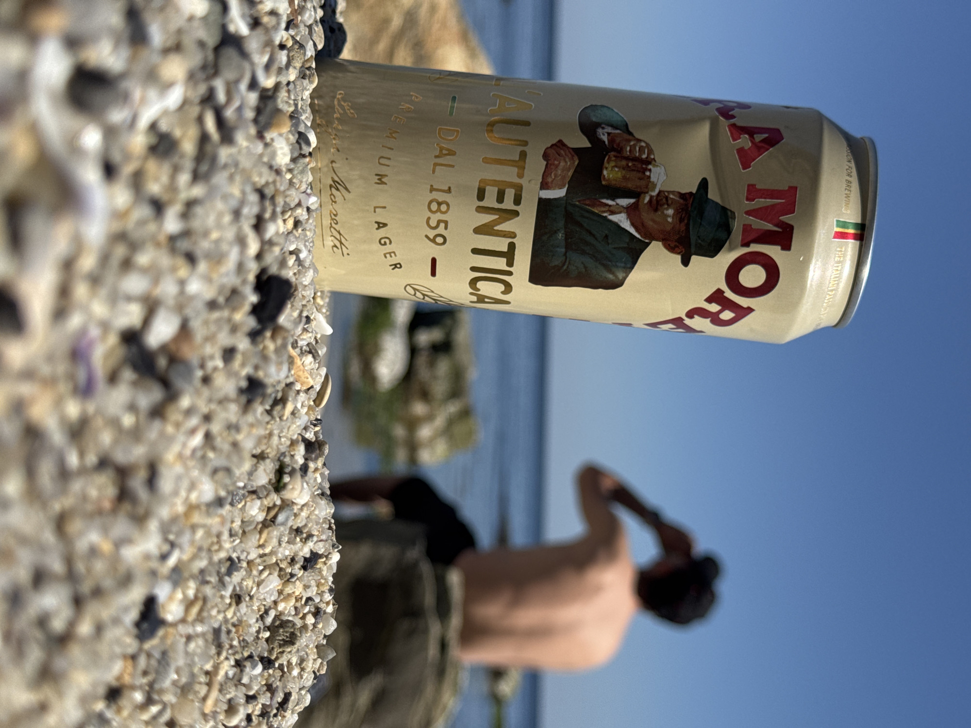 The image shows a can of La Moretti L'Autentica premium lager beer resting on a pebble beach. The label includes the text "DAL 1859" and "Luigi Moretti." In the background, a person with their shirt off is standing near the water. The sky is clear and blue. The specific location in Varna, Bulgaria is not discernible from the image.