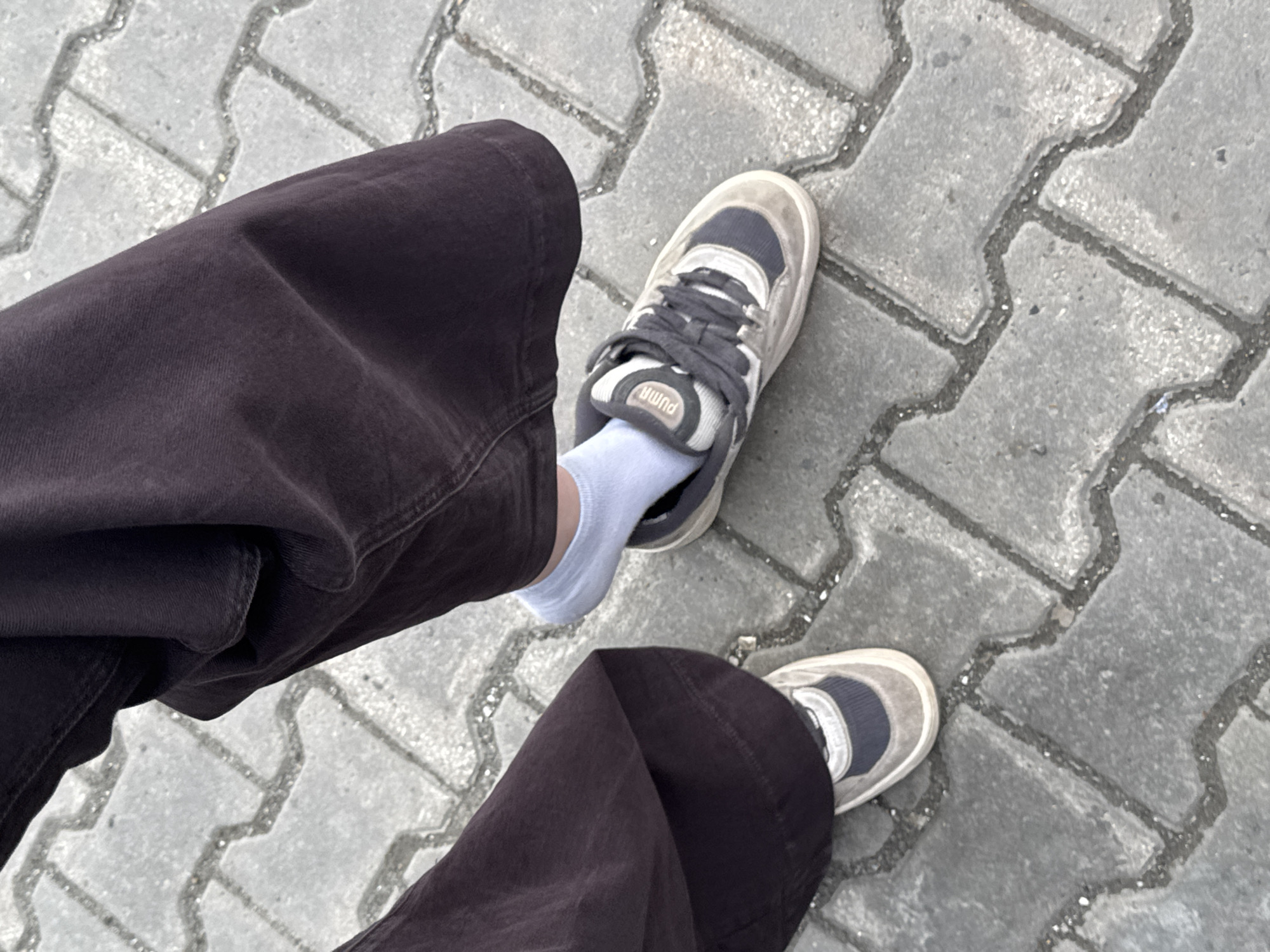 The image presents a top-down view of a person's legs and feet standing on grey interlocking paver stones. The individual is wearing dark, possibly wide-leg, pants, which are wrinkled and folded around the ankles. Light blue ankle socks are visible above the sneakers. The footwear consists of a pair of sneakers, predominantly light grey or beige with darker grey accents and laces. The brand "PUMA" is clearly visible in white text on the tongue of the right shoe.
The ground is paved with patterned concrete or asphalt blocks, featuring a wavy, interlocking design with dark grout lines between them, typical of pedestrian areas in urban settings like Perm, Russia. The lighting suggests it is daytime, with no shadows indicating a specific time of day or harsh sunlight. The scene depicts a momentary pause, possibly mid-stride, showcasing the person's casual attire against an outdoor backdrop. No other subjects or events are visible, and no specific emotions can be inferred from the perspective.