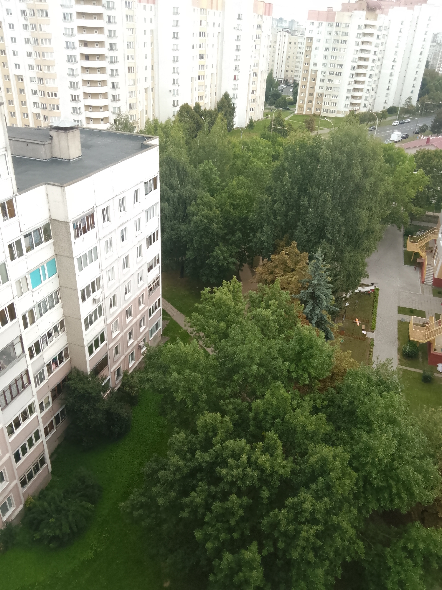 An aerial view of a residential area in Minsk, Belarus. Multiple tall, multi-story apartment buildings, predominantly white with cream and pink accents, dominate the scene. Lush green trees and foliage are interspersed between the buildings, with some appearing to be within courtyards or along pathways. A grey paved walkway is visible, leading towards one of the buildings. A small playground area with orange and white structures is also present in the scene, as are several parked cars on a road in the distance. The sky appears overcast.