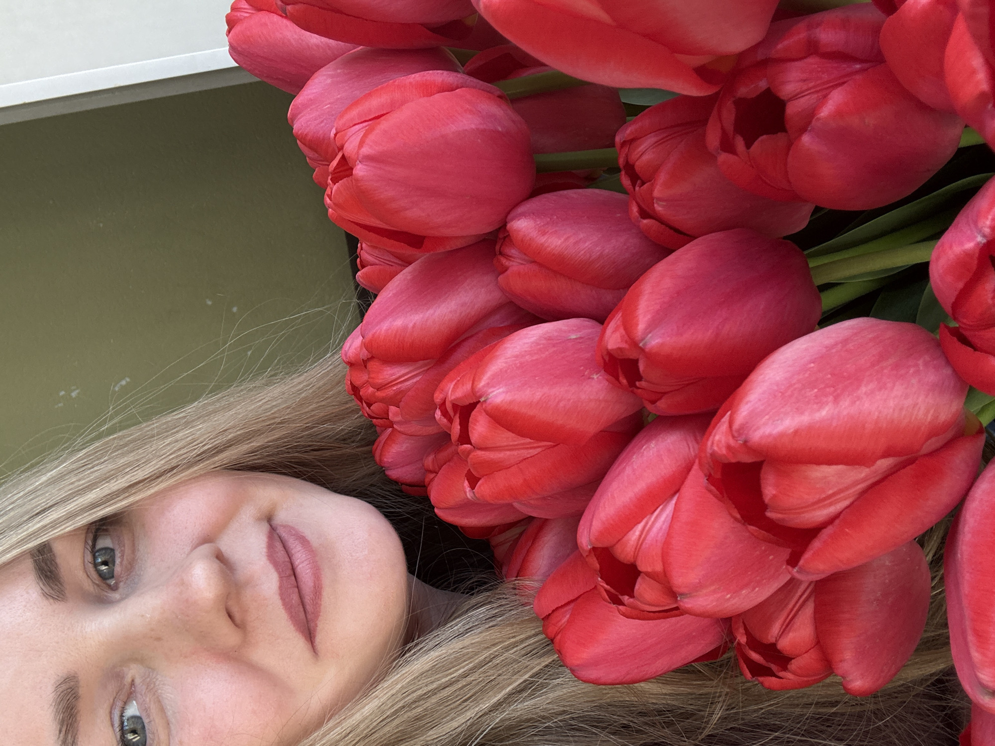 A woman with blonde hair is next to a large bouquet of red tulips. The background includes a wall, possibly indoors. The exact location within Chaikovsky, Russia, cannot be determined from this image alone.