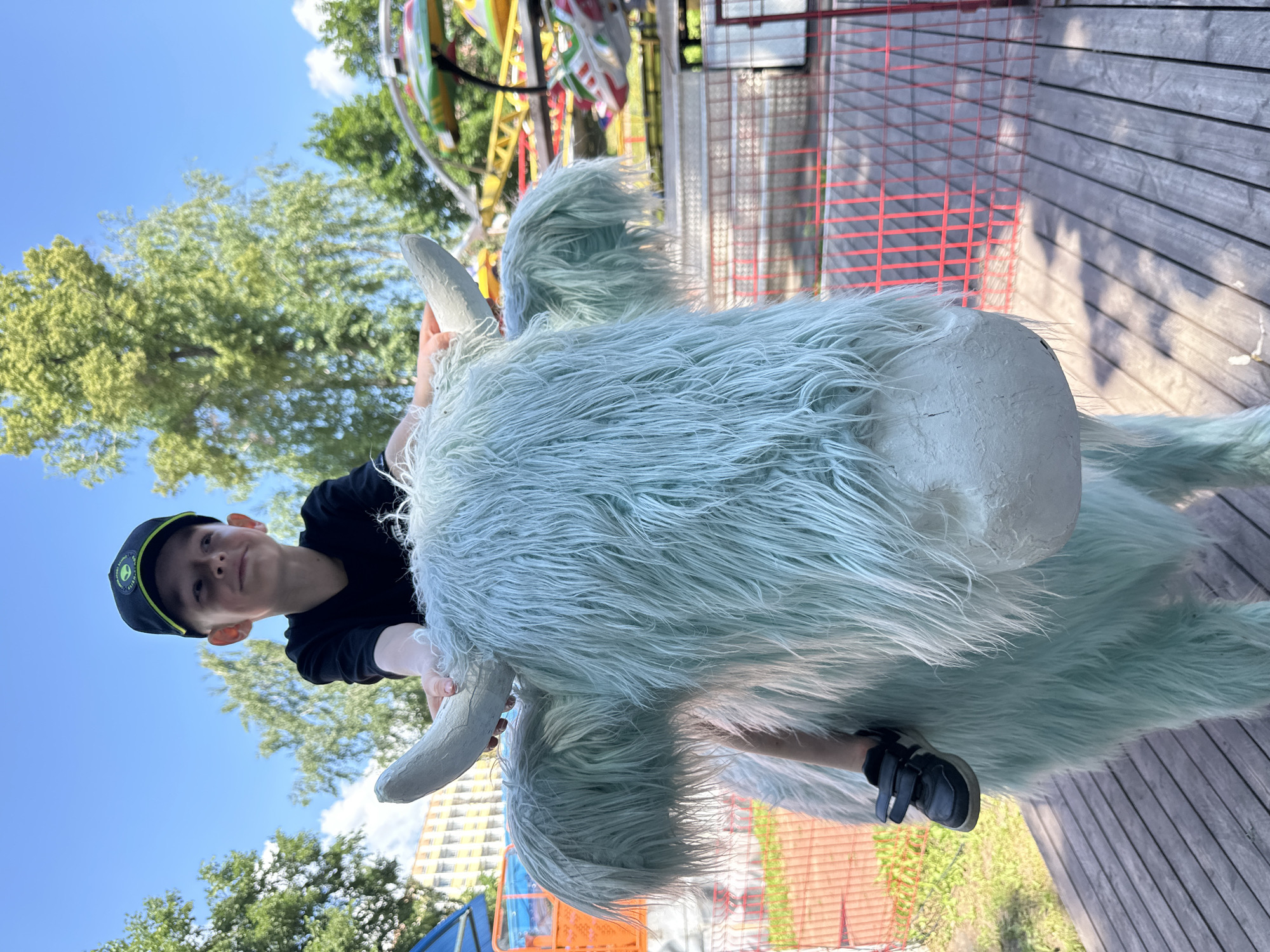 The image captures a young boy, wearing a black t-shirt and a black cap with an alien emblem, sitting atop a large, fuzzy, light blue artificial yak-like creature. The boy is smiling and looking to his left, with his hands resting on the creature's horns. His legs are dangling on the side. The creature has long, shaggy fur and pale, large horns.
The scene appears to be an outdoor amusement area, likely a park or a fairground, as suggested by the presence of a colorful amusement ride in the background, featuring yellow and red structures. A red-fenced area is visible, separating the amusement ride from the wooden platform where the yak and the boy are situated. Green foliage and trees are present in the background, under a clear blue sky with a few scattered clouds. The ground is covered with a wooden plank walkway. Based on the context of similar images, this scene is likely located at the Udmurt State Circus in Izhevsk, Russia, which is known to have a children's entertainment zone that includes various attractions like this one.