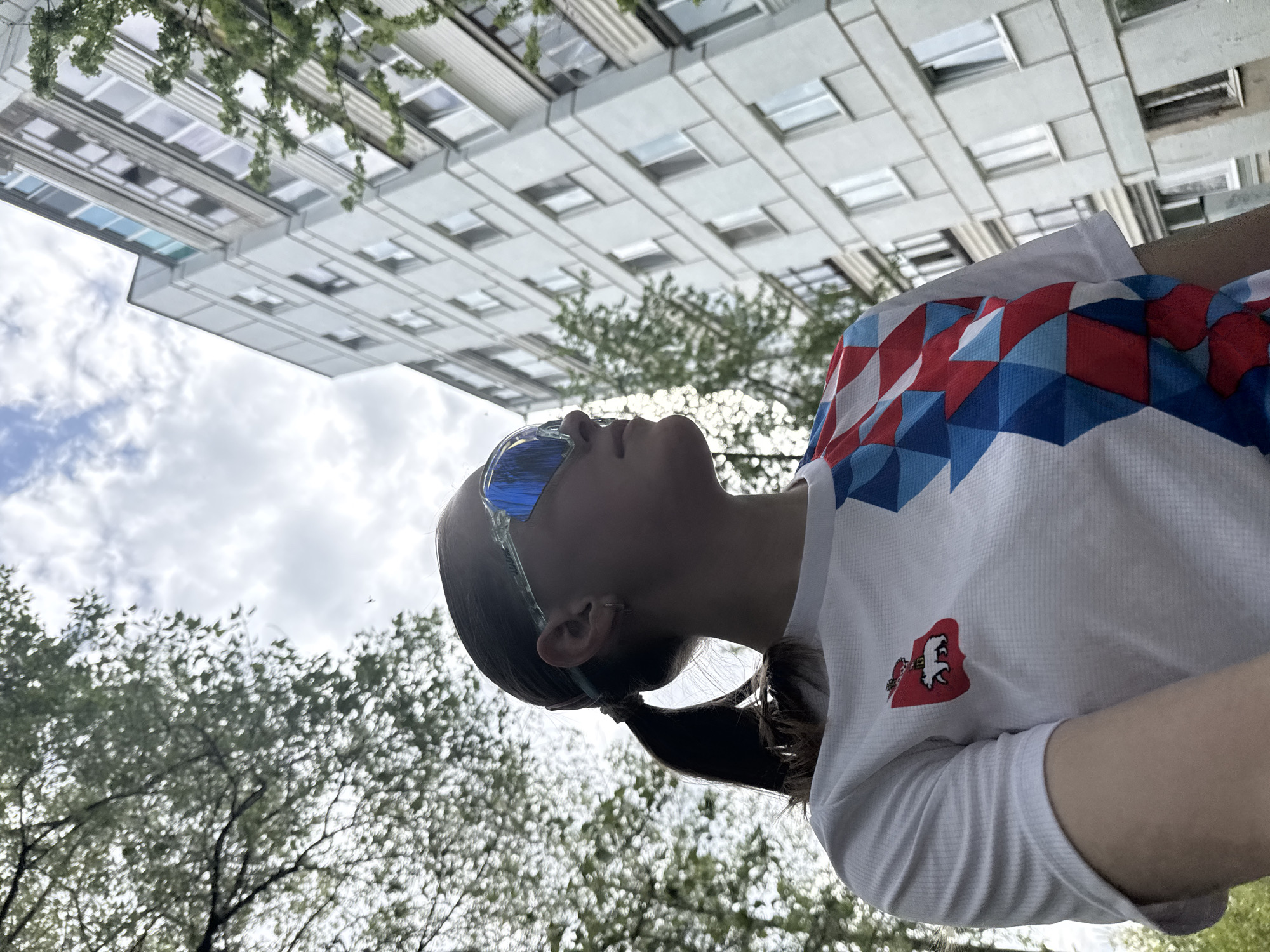 A young woman with a ponytail is positioned in the lower half of the frame, gazing upwards at a building in the city of Chaikovsky, Russia. She is wearing mirrored sunglasses and a white t-shirt featuring a pattern of red and blue geometric shapes, along with a distinct coat-of-arms emblem on the lower left. The t-shirt's design suggests a possible connection to a sporting event or national team. The background includes a multi-story building with numerous windows, partially obscured by the branches and leaves of trees. The sky is visible through the tree foliage, with patches of blue and white clouds. The photograph is taken from a low-angle perspective, giving the impression of looking up at the woman and the surrounding environment.