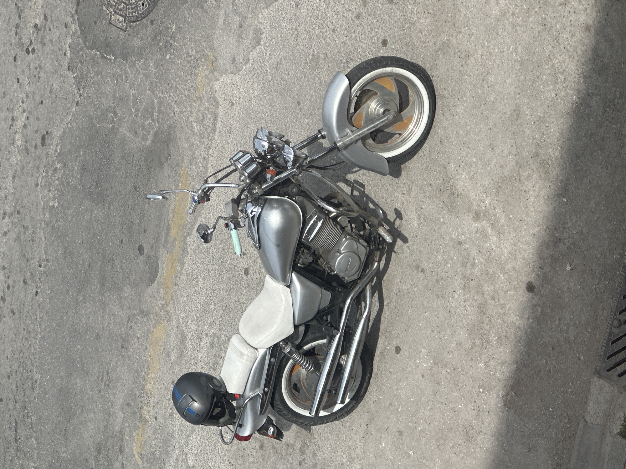 Observed from above, a silver motorcycle with white tires is parked on a gray concrete surface. A dark helmet rests near the motorcycle's rear. The location appears to be an outdoor paved area. A manhole cover is visible in the upper left corner. It appears to be daytime, with sunlight casting shadows. Based on the provided information, the location is the city of Fethiye, Turkey.