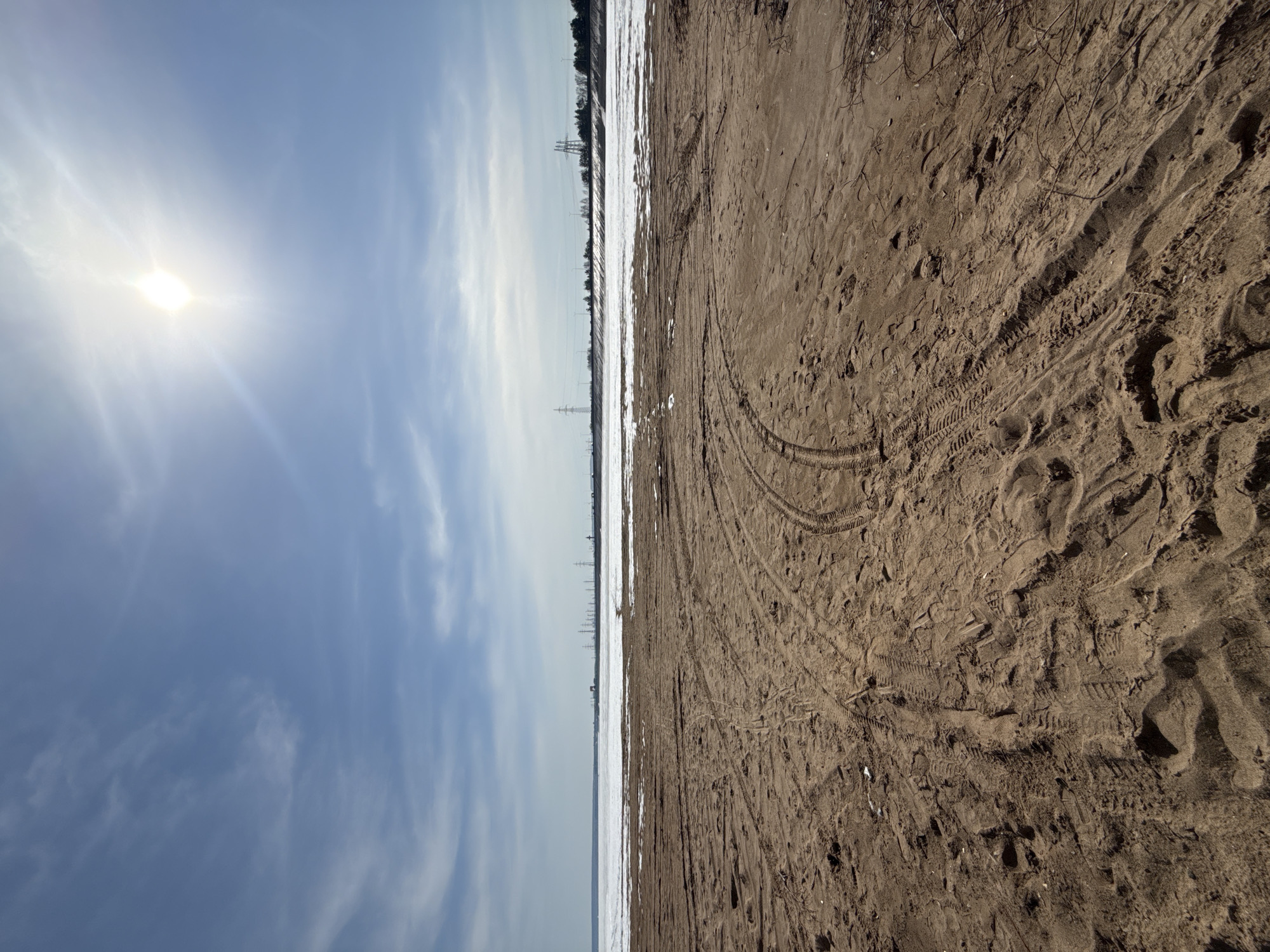 The image depicts a horizontal division of contrasting landscapes. The left side showcases a bright sky with light clouds and the sun. The right side features a sandy or earthen ground covered in tracks, possibly tire marks or footprints. A strip of land or a shoreline with remnants of snow or ice separates the two halves. In the background, utility poles and trees are visible, suggesting a landscape near an industrial or residential area.
Based on the prompt's mention of Noviy, Russia, the image may depict a scene near a body of water or a snow-covered field in the area. The photo suggests a cold season, with the remnants of snow and a bright but possibly cold sun.