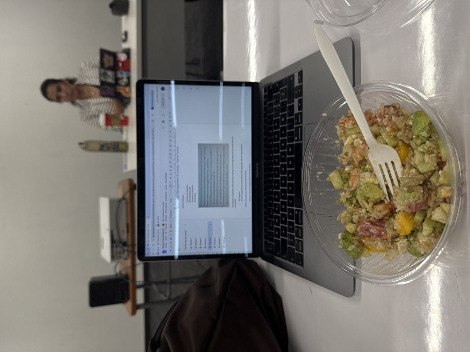 An indoor scene is depicted from a low-angle, first-person perspective. In the immediate foreground, a clear plastic bowl holds a salad containing quinoa, diced avocado or cucumber, and orange-colored fruit, possibly mango. A white plastic fork is partially submerged in the salad. To the right, a clear plastic lid rests on a white tabletop.
Directly behind the salad, a silver MacBook laptop is open on the same white tabletop. Its screen displays a document within a word processing application, titled "Project Management (5 points) Diana Cortes," featuring a list of activities. To the left of the laptop, a dark brown bag is partially visible.
In the mid-ground, a woman with dark hair, wearing a light-colored striped blazer over a dark garment, is seated at a white table, positioned out of focus. She appears to be looking towards the left side of the frame. On her table are a tall, slender metallic bottle and a red and white disposable coffee cup. A dark device, possibly a laptop or tablet, adorned with colorful elements, is situated in front of her.
Further to the left, a black speaker and a white multimedia projector are visible on a small wooden table or stool. Cables are connected to the projector. The background consists of plain white walls and a dark floor. The specific location within Mexico City, Mexico, cannot be determined from the image.