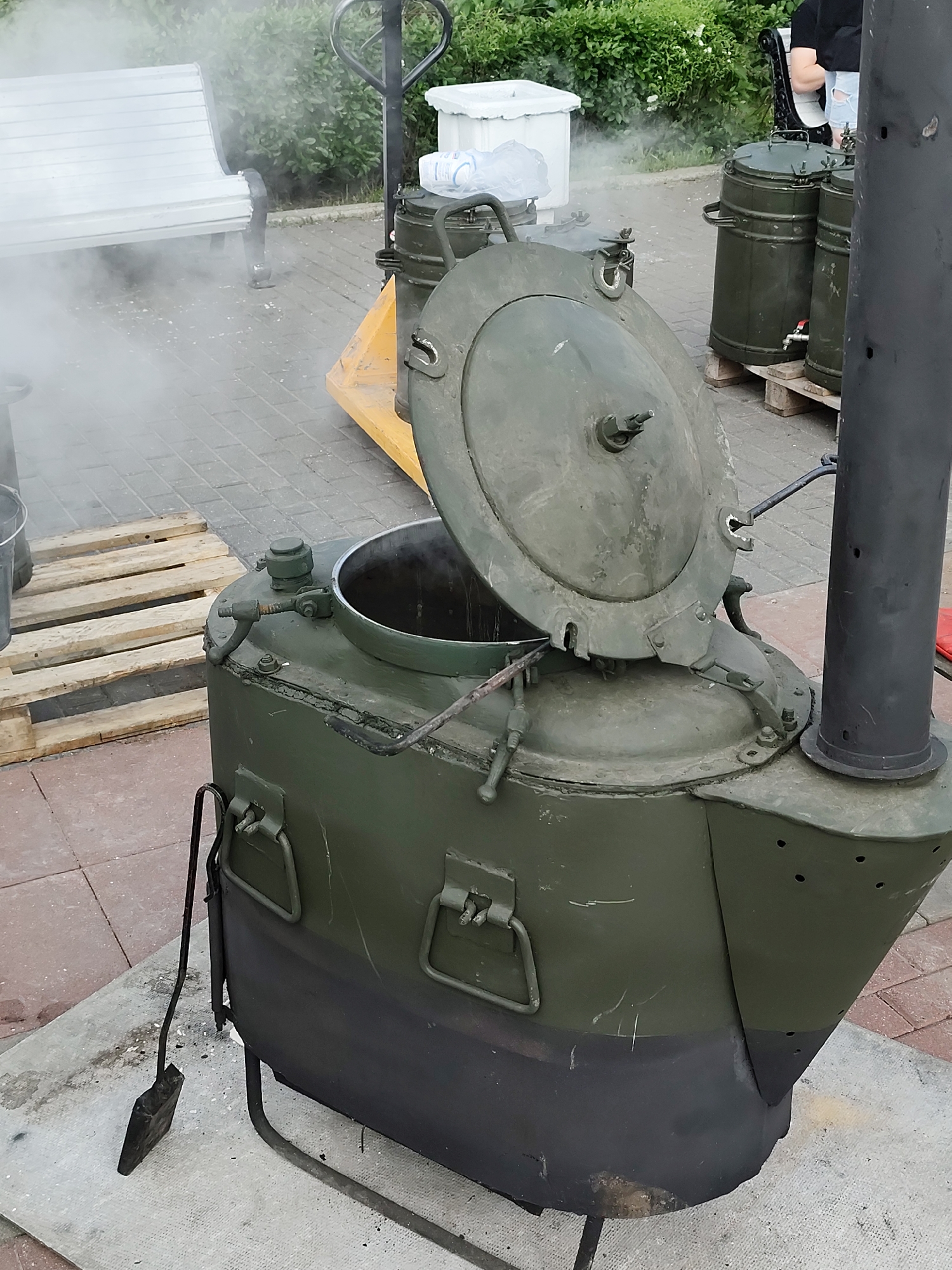 The image shows a large, olive-green military field kitchen cooker, common in former Soviet countries. The lid of the cooker is open, and steam is rising from inside. The cooker is mounted on a black base with thin metal legs, and a black chimney is attached to the side. A metal shovel leans against the side of the cooker.
The cooker is located on a paved surface, possibly a sidewalk or courtyard, with wooden pallets nearby. In the background, there are other similar cookers, a hand truck, a white bench, and some foliage. The location could be a park or public space in a city. Given the prevalence of such cookers in former Soviet countries, and the style of the bench, the location might be a park in Moscow, Russia.
