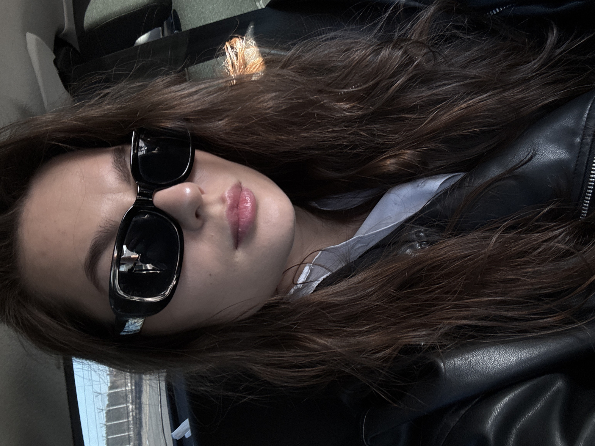 An eye-level close-up shot captures a young person, likely female, inside what appears to be a vehicle in Chaikovsky, Russia. The image is rotated counter-clockwise, showing the subject's head tilted to the right. She has long, wavy dark brown hair that cascades around her shoulders and chest.
The subject wears black rectangular sunglasses that cover her eyes, reflecting what seems to be the vehicle's interior. Her lips are slightly parted, showing a light pink lip color. She is dressed in a black leather jacket over a white collared shirt, the collar peeking out from beneath her hair.
In the background, elements of the vehicle's interior are visible, including a dark headrest towards the top. On the left side of the image, a window shows a blurred view or reflection of what appears to be urban buildings or structures, indicating an outdoor setting beyond the vehicle. The lighting suggests it is daytime. The person is posing for the photograph, possibly a selfie, with a neutral or slightly pensive expression. No text is visible in the image.