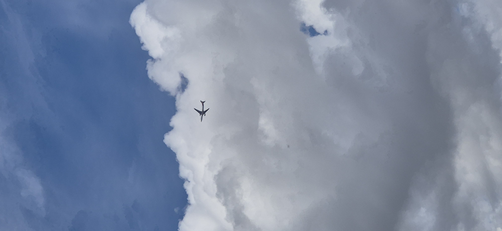 A commercial airplane is captured in mid-flight against a backdrop of a cloudy blue sky. The aircraft, silhouetted against the clouds, is positioned in the upper central portion of the frame, appearing to be at a considerable altitude. The clouds are varied, with both bright white, puffy formations and darker, more diffused grey masses. The overall scene suggests a daytime view from below the aircraft. The location in the city of Mainz, Germany, is not discernible from the visual information provided.