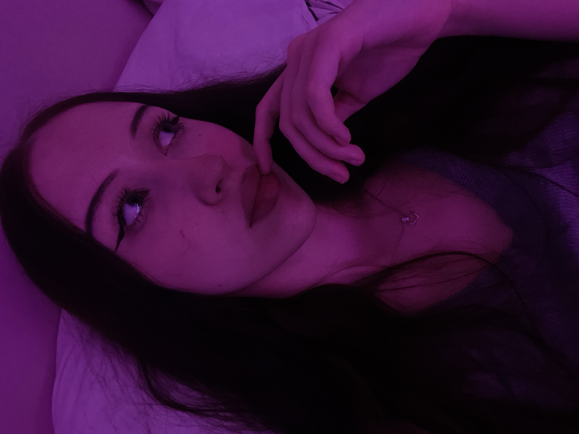 The image shows a close-up, overhead, dimly lit selfie of a young woman lying on a bed. The room is illuminated by purple lighting, casting a strong magenta hue over the scene. The woman has long, dark hair spread out around her head and shoulders. She is wearing a grey top and a small silver heart-shaped pendant necklace. Her right hand is raised, with her index and middle fingers touching her lips. Her eyes are looking to her right, and she has winged eyeliner on. The background consists of a white pillow and bedding, also illuminated by the purple light. There is no visible indication of the location being Votkinsk, Russia.