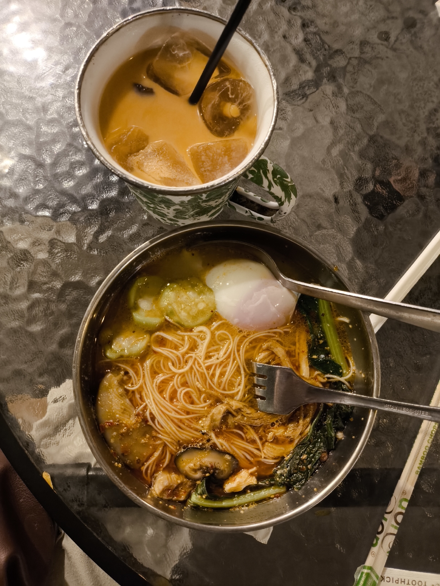 A top-down view shows a glass-topped table with two dishes of food and a drink. To the left, a white mug with green and white patterned detail contains a beverage with ice cubes and a black straw. To the right, a metal bowl holds a dish of noodles in broth, topped with a poached egg, sliced cucumbers, mushrooms, and leafy greens. A spoon and fork are resting on the rim of the bowl. To the right of the bowl, a pair of chopsticks in a wrapper are visible. The reflective surface of the table suggests it is made of glass. The presence of a beverage and noodle dish implies a cafe or restaurant setting. While the image does not explicitly state the location, such a scene is common in food establishments in cities like Surakarta, Indonesia, known for its diverse culinary offerings.