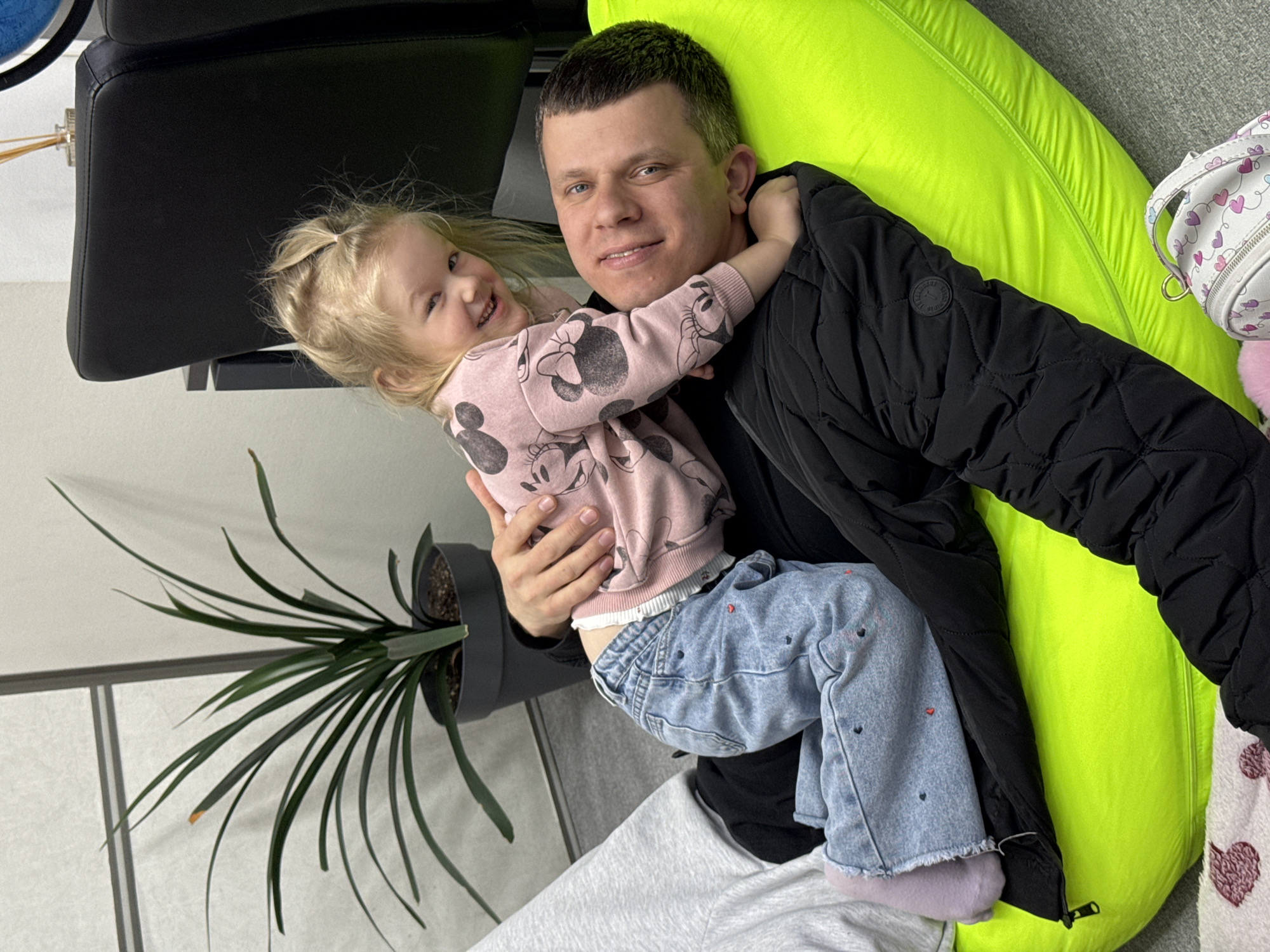 The image, captured in Perm, Russia, depicts a joyful indoor scene featuring an adult man and a young child. The man, with short dark hair and light eyes, is smiling warmly at the camera, his arm around the child. He is wearing a black quilted jacket with a visible circular patch on the left arm that reads "D SQUARE DENIM," and light grey sweatpants are partially visible at the bottom.
He is holding a blonde-haired child, likely a girl, who is also smiling widely and looking at the camera. Her arms are affectionately wrapped around the man's neck, suggesting a close bond. The child is dressed in a pink long-sleeved top featuring multiple black Mickey Mouse head silhouettes and light blue jeans adorned with small red heart patterns. Her feet are covered in light purple socks or soft shoe linings.
The setting is a brightly lit indoor space, possibly a living area. A vibrant neon green beanbag chair or cushion is behind the man and extends to the right. A black leather chair is partially visible in the upper left background. In the lower left, a potted plant with long, thin leaves adds a natural element. The walls or floor appear to be light-colored, with some shadows indicating natural light. The expressions of both individuals convey happiness and affection, creating a heartwarming atmosphere.