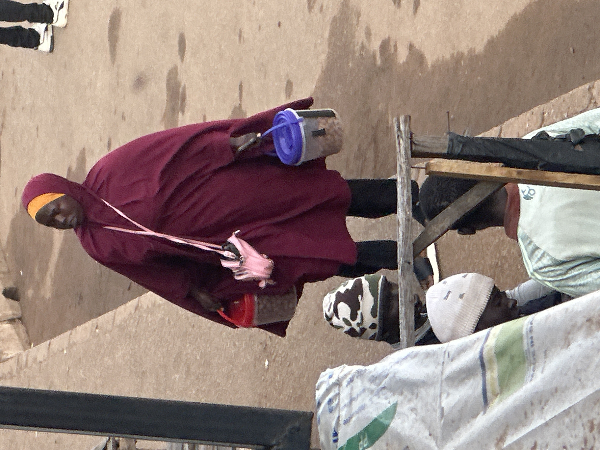 In Bukuru, Nigeria, a person wearing a burgundy hijab and a yellow headscarf stands in the center of the frame, carrying two buckets by their handles. The bucket on their left is red with a clear plastic container and contains what appears to be food items. The bucket on their right is blue with a clear plastic container and also holds food items. A pink clutch bag is slung over the person's shoulder. In the foreground, three other individuals are partially visible. One person on the left is wearing a white and blue patterned garment. Another individual in the center is wearing a camouflage-patterned hat. To the right of that person, another individual is wearing a white knitted cap. The ground is a light brown, dusty surface. A metal railing with a wooden plank runs horizontally in the foreground.