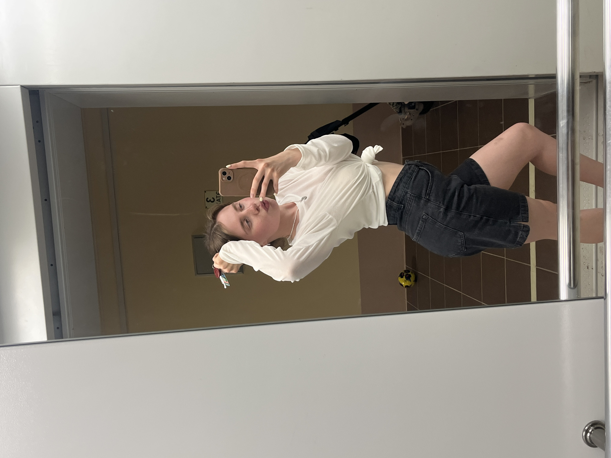 The image is a mirror selfie featuring a young adult female in an indoor setting within Chaikovsky, Russia.
The woman, with light brown hair, is wearing a white long-sleeved crop top tied at the waist, exposing her midriff, and dark charcoal denim shorts. She has light-colored nail polish and a silver necklace with a small pendant. Her left arm is raised with a set of keys, featuring red and white fobs, held near her head and intertwined with her hair. Her right hand holds a light brown-cased smartphone to capture the image, and she is posing with pouted lips.
The reflection in the mirror reveals a room with light brown/tan walls and a brown tiled floor with darker grout lines. A small, yellow and black patterned ball rests on the floor to the left of the woman's reflected figure. Partially visible behind her reflected right side is a dark-colored object resembling part of a baby stroller or wheeled equipment. Above her reflected head, on the far wall, a reversed sign displays characters that appear to be a stylized '3' followed by letters resembling 'EATS' or 'КАТЕ' in Cyrillic. A horizontal white or light-colored line is visible across the tiled floor behind her. The mirror itself is framed by white architectural elements, and a silver-colored metal handrail is present in the foreground at the bottom of the frame. The lighting in the room appears to be overhead, providing general illumination.