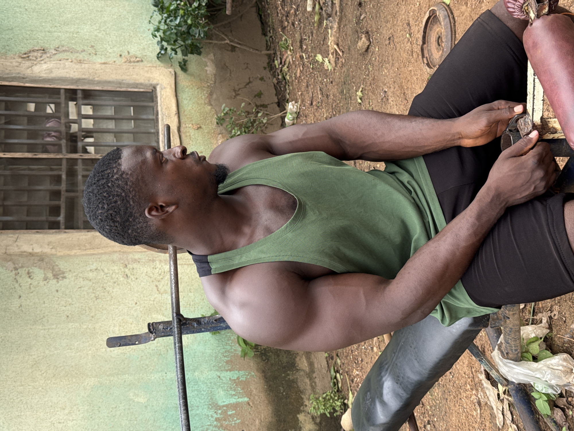 In Akwanga, Nigeria, a muscular Black man with short, dark hair and a goatee sits on a black weight bench outdoors. He is wearing a green tank top and black shorts. The bench is positioned in front of a light green, weathered wall with a barred window. The ground is covered in dirt and fallen leaves, with some green plants growing near the bench. A metal barbell rack is visible behind him, and a weight plate is on the ground to his right. He is looking to his right and appears to be holding onto a part of the bench with his hands.