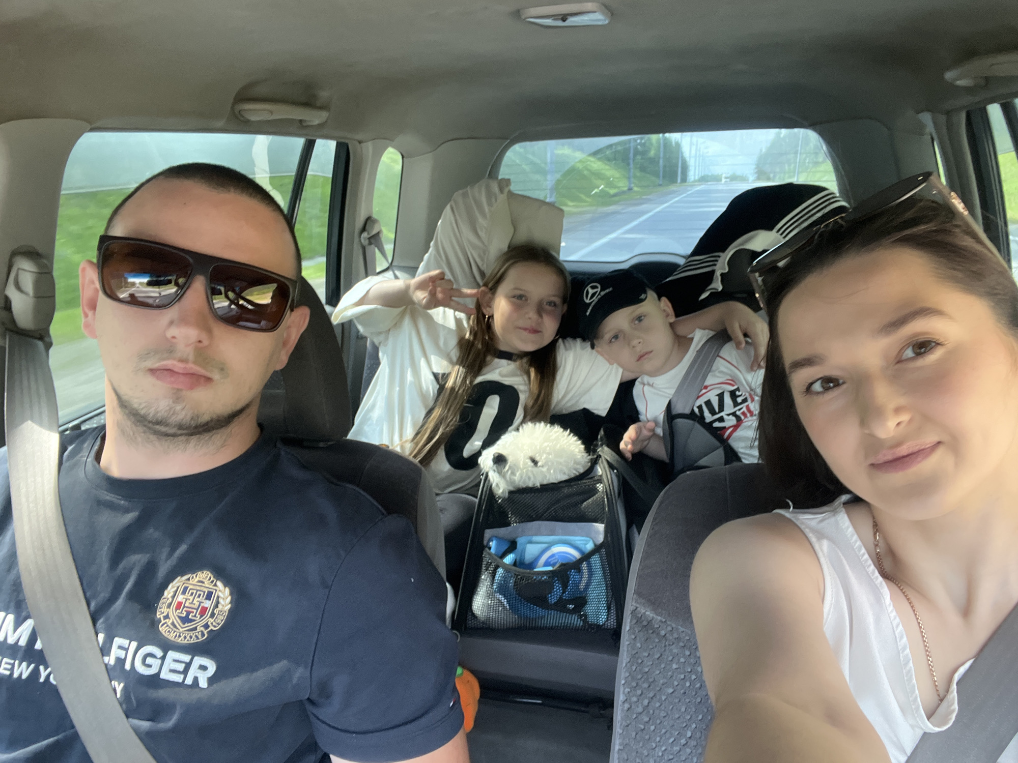 A family of four, including two adults and two children, are traveling in a car. The adult male in the front passenger seat wears sunglasses and a navy blue Tommy Hilfiger t-shirt. A white dog sits in a carrier in the back seat between the two children. The adult female in the rear passenger seat has dark hair and is wearing a white top. The children are a girl and a boy. The girl is making a peace sign with her fingers. They are on a road trip, possibly in the city of Novolisikha, Russia, with green fields and trees visible outside the car windows. The car appears to be an SUV, and the interior is visible, including car seats and other passenger accessories.