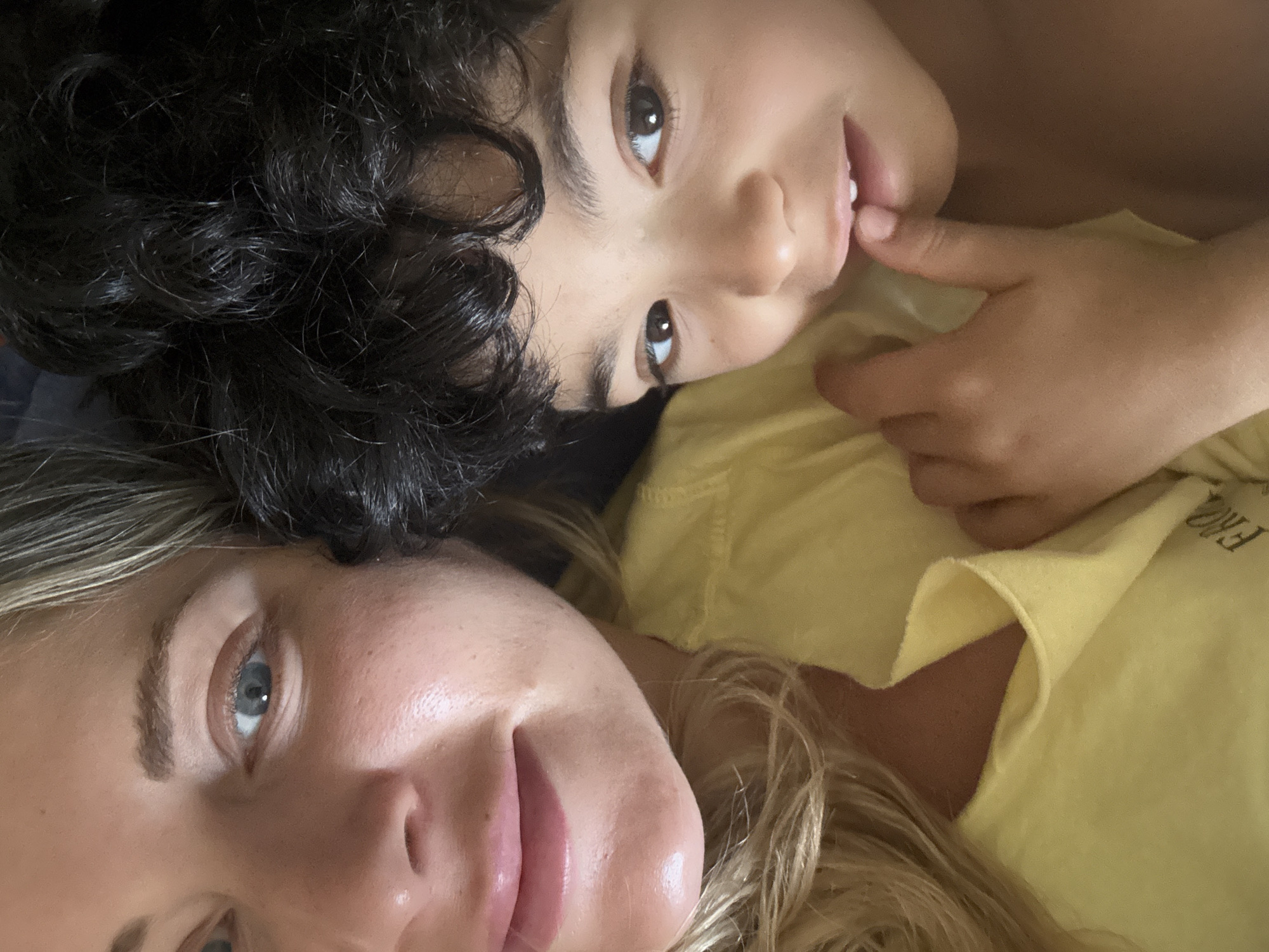 Two people are in the image. A fair-skinned woman with blue eyes and blonde hair is at the bottom left of the frame. A child with dark curly hair and brown eyes is at the top right, with a finger touching their lip. Both are looking towards the camera. The child is wearing a yellow shirt, as is the woman. The location is unknown.