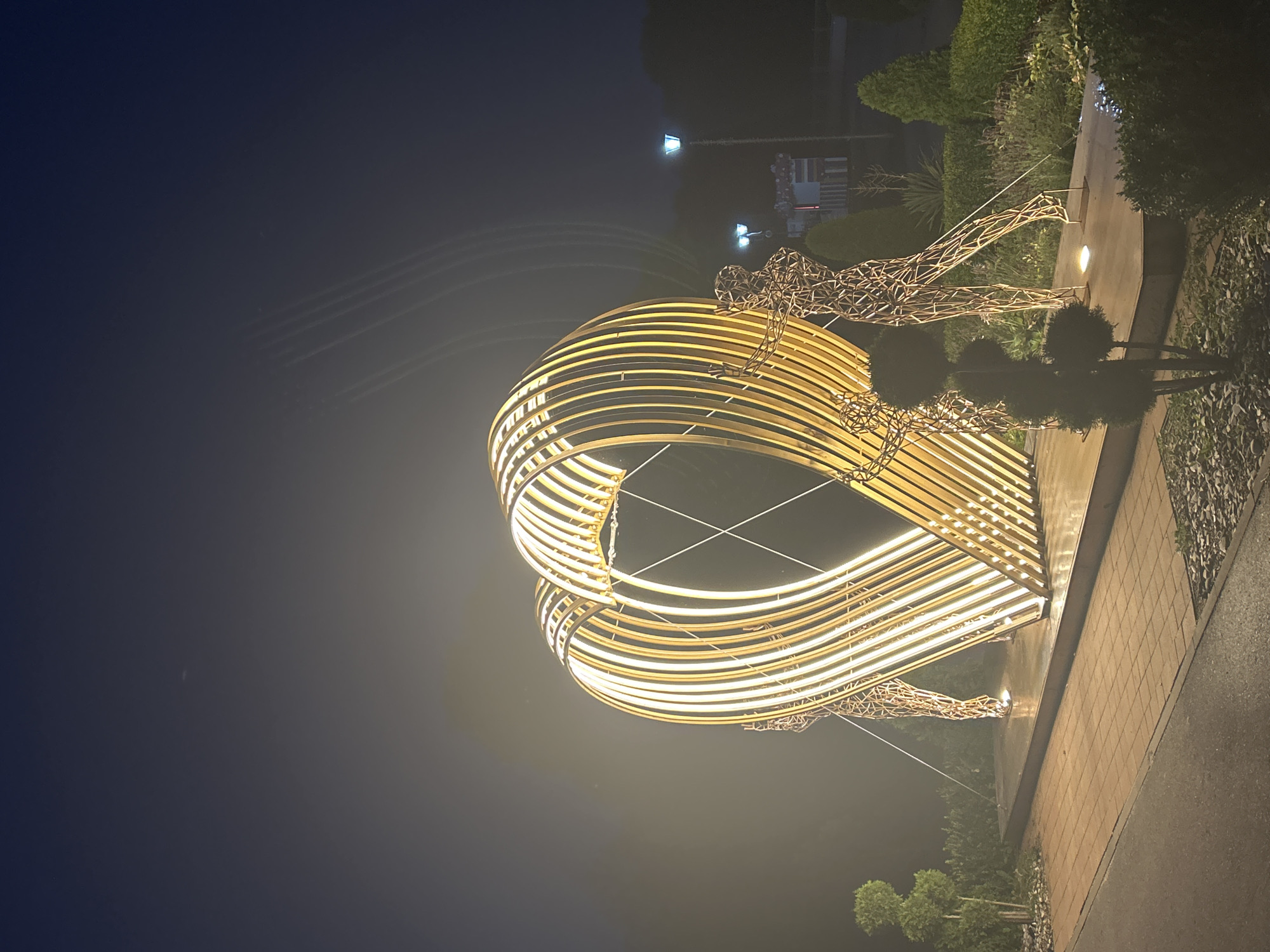 In Brest, Belarus, at night, a large, illuminated heart-shaped sculpture stands as the central focus. The sculpture is composed of numerous vertical golden-colored slats, creating a cage-like effect that outlines the heart's form. The interior of the heart is lit by warm yellow lights. Two wireframe sculptures of human figures are positioned at the base of the heart sculpture, appearing to hold or support it. One figure is on the left and the other on the right. The scene is set in an outdoor landscaped area with paved pathways, gravel beds, and various types of bushes and trees, including topiaries. Streetlights with blue-toned bulbs illuminate the background. The sky is dark, suggesting nighttime, with faint light trails visible in the upper portion of the image. The overall impression is a well-lit public art installation in a park-like setting.