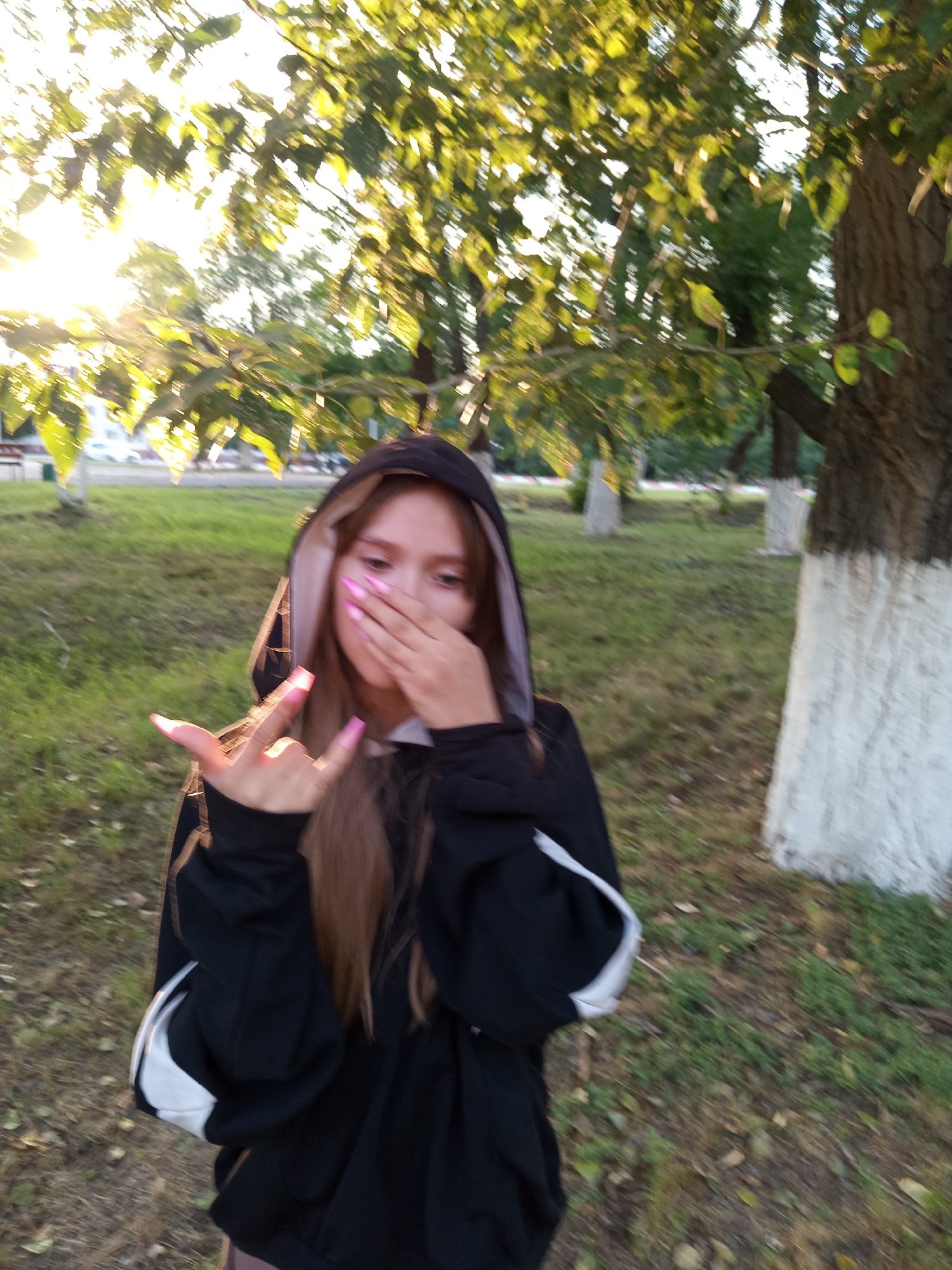 A young woman with long brown hair, wearing a black hooded sweatshirt with white accents, stands outdoors in a park-like setting. She is covering her mouth with her right hand, her left hand is raised with her index and pinky fingers extended, and she has bright pink fingernails. The background consists of grass, trees, and what appear to be painted tree trunks. The lighting suggests it is either sunrise or sunset. The location is likely a public park in the city of Rudny, Kazakhstan.