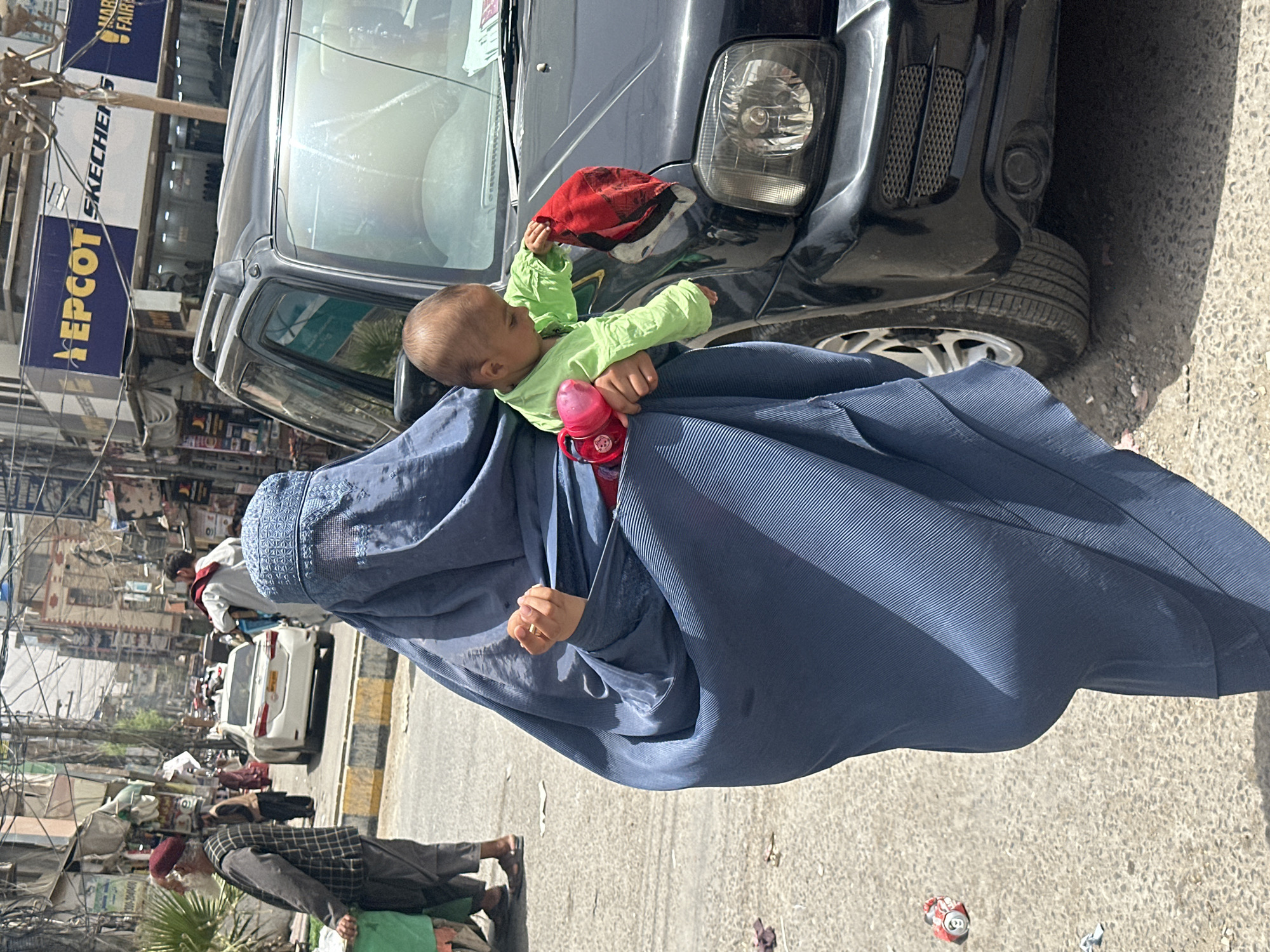 A woman in a blue burqa holds a baby in a green shirt. The baby is holding a red bottle and touching a red cap on the hood of a black vehicle. The woman is wearing a gold ring on her finger. The scene is on a city street in Quetta City Tehsil, Pakistan. A man walks by in the background, and there are shops and cars visible on the street. A Skechers sign is present. A discarded soda can lies on the street.