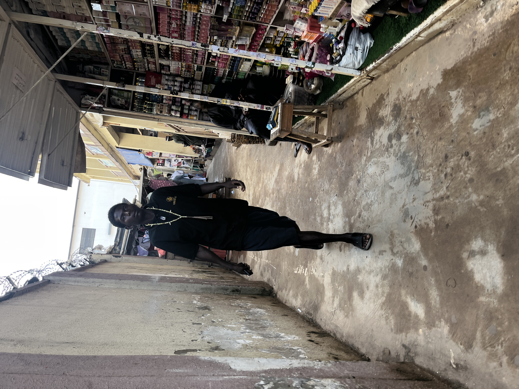 A man in a black t-shirt and blue shorts walks down a narrow, muddy alleyway in Abuja, Nigeria, on the left side of the frame. He wears a beaded necklace and sandals. To his right, a stall is filled with various products, primarily bottles and containers, arranged on shelves. A man in black attire sits on a stool in front of the stall, appearing to be attending to business. The ground is wet and uneven, with evidence of standing water. A textured wall with peeling paint runs along the left side of the alley, topped with barbed wire. The background shows buildings and other stalls, suggesting a market environment.