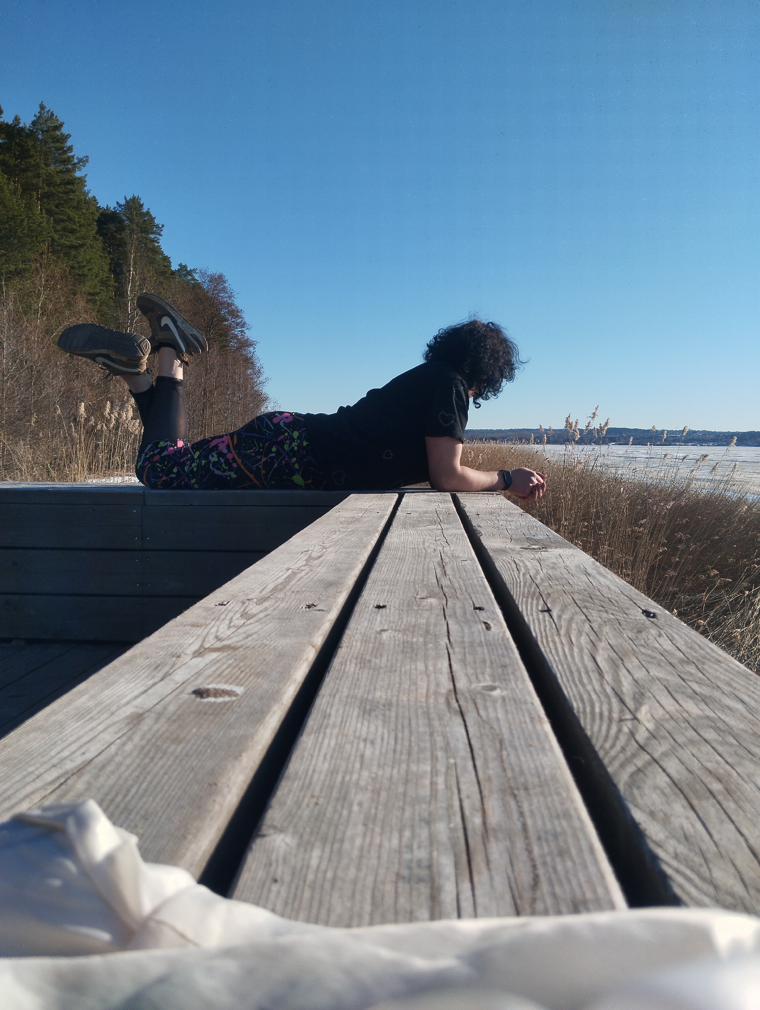 A person is lying on a wooden platform overlooking a body of water, possibly a lake or river, in Votkinsk, Russia. The person is wearing black leggings with a multicolored splatter pattern and dark athletic shoes. They also have a black short-sleeved shirt. The person is resting on their elbows, looking out at the landscape. The water appears to be partially frozen, with reeds or tall grasses growing along the shoreline. A forested area is visible in the background. The sky is clear and blue. A white object is visible in the immediate foreground.