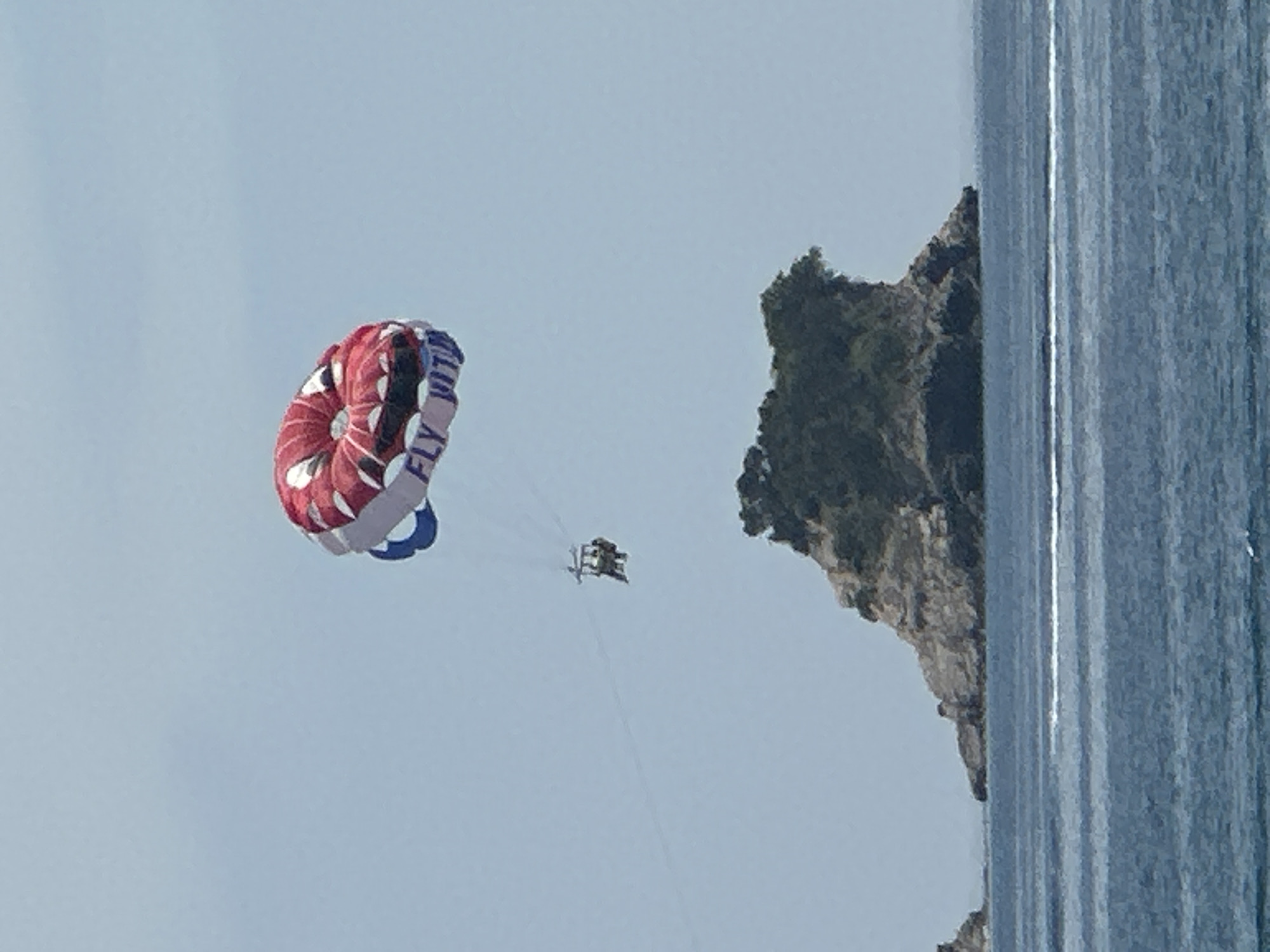 The image shows a bright, clear day over a body of water with a rocky island in the distance. A parasailer, with a red and white parachute with blue accents, is visible in the sky, appearing to be pulled by a boat not visible in the frame. Two people are seated in the harness beneath the parachute. The rocky island is covered in dark green vegetation and rises out of the blue water. The scene appears to be a popular tourist destination, possibly the coast of Budva, Montenegro, known for its beautiful scenery and water sports. The water is a deep blue, with some whitecaps indicating movement. The sky is a pale blue, with no clouds visible.