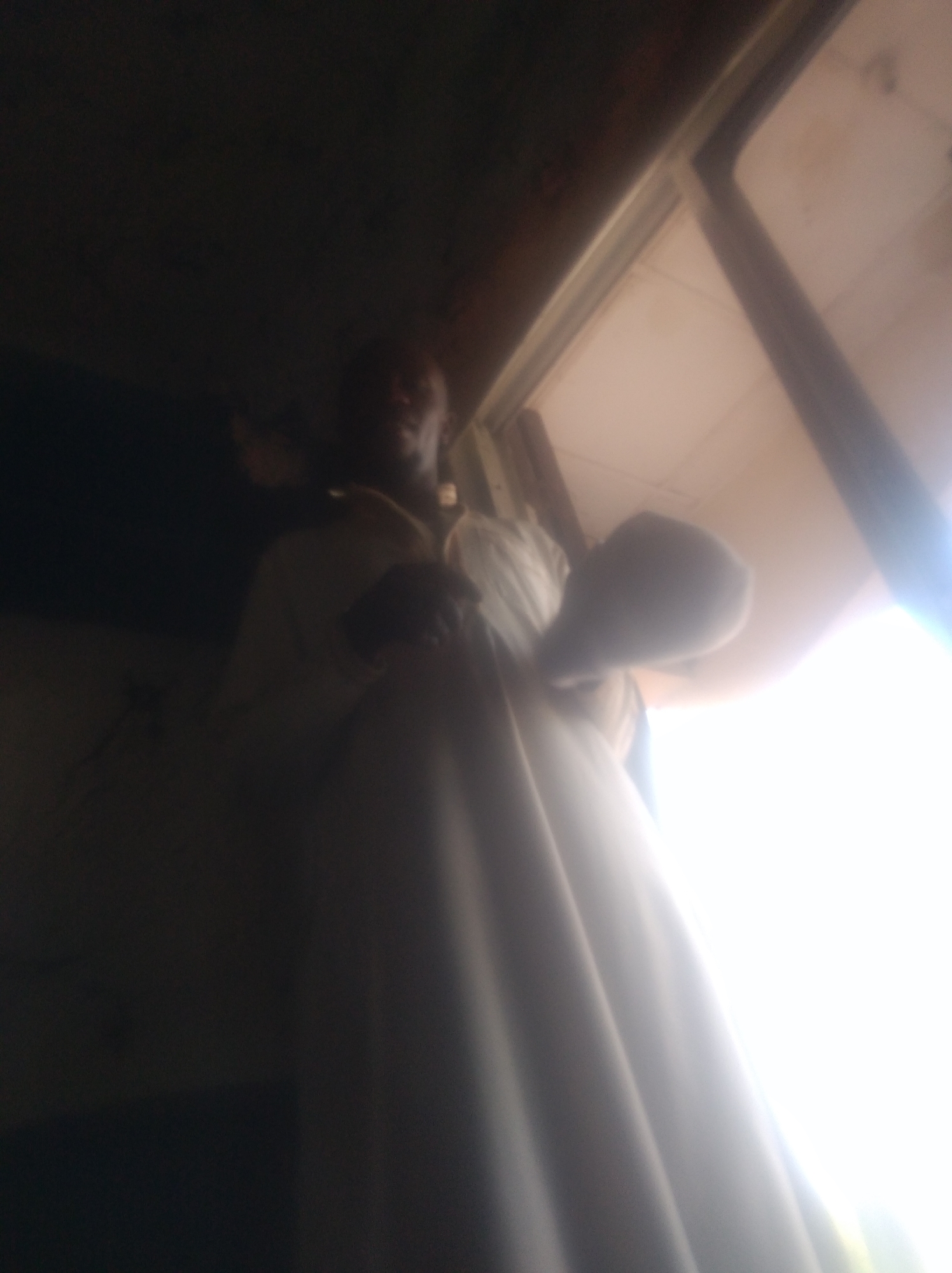 This is a vertically oriented, low-angle, blurry photograph taken indoors, likely during the daytime given the bright light emanating from a window. The primary subject appears to be a person wearing a white garment, possibly a religious robe or traditional attire. The person is partially obscured by shadows and the low resolution of the image, but their face is visible, looking towards the camera. Their skin tone suggests they are of African descent.
The person is standing near a window, with the bright light from outside creating a strong contrast with the darker interior. The light reflects off the white garment, making it appear luminous. A portion of what looks like a ceiling is visible at the top of the frame, and a dark, textured surface to the left might be a wall or furniture. The overall setting is domestic or religious.
The activity seems to be a moment of pause or presentation, with the subject positioned prominently in relation to the light source. The mood is somewhat solemn or introspective, conveyed by the shadowed features and the stark lighting. No text is visible in the image. The image is dated to Jalingo, Nigeria, suggesting a specific geographic and cultural context.