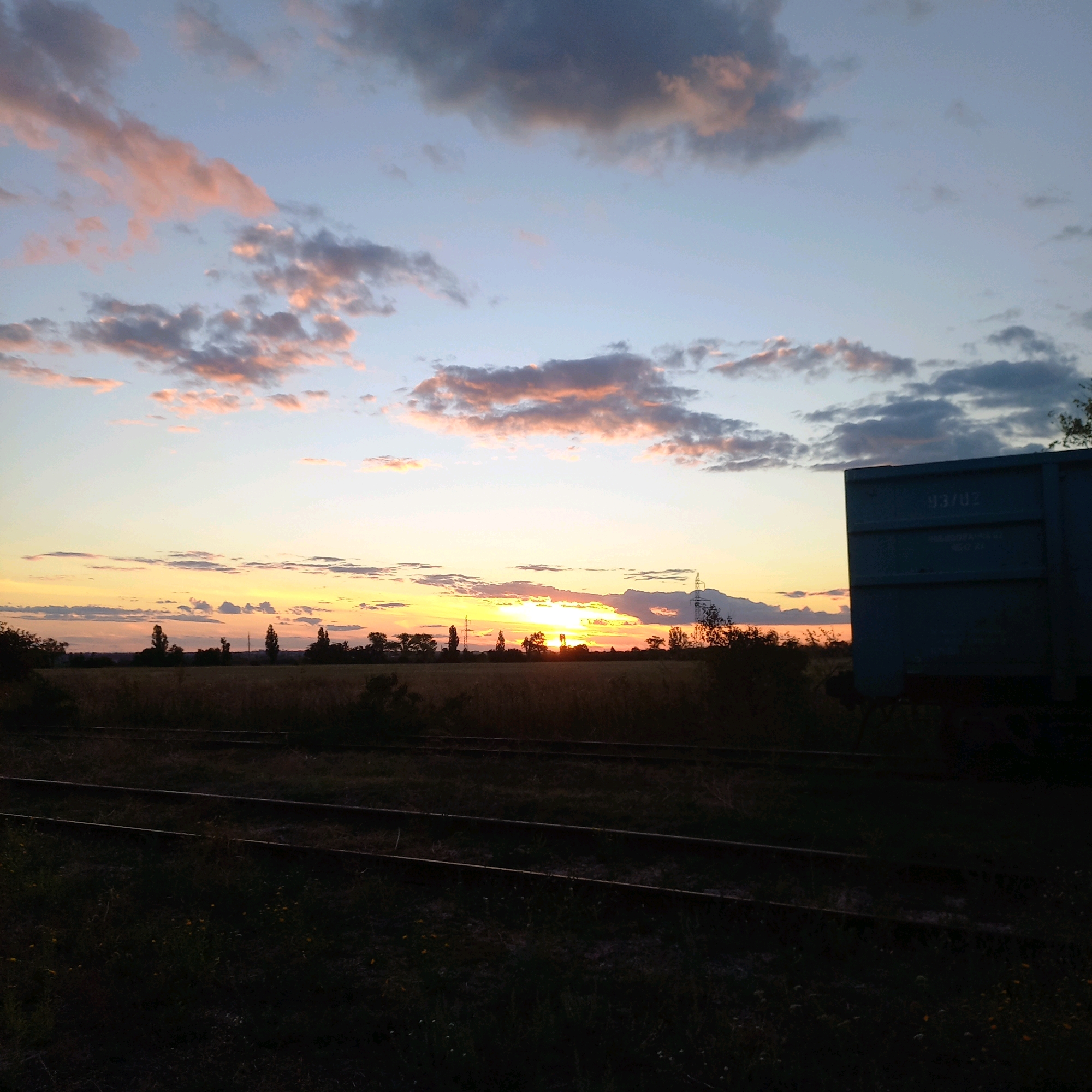 A wide-angle, eye-level, outdoor photograph captures a sunset over a rural landscape with railroad tracks in the foreground. The sky is filled with wispy clouds tinged with pink and orange, transitioning to a pale blue. The sun is low on the horizon, casting a warm glow across the scene. In the middle ground, a flat, dry field is dotted with silhouetted trees and a few power poles. A blue freight car is visible on the right side of the frame, partially obscuring the field. The foreground features a set of railroad tracks running horizontally across the image, with dark grass and some small yellow flowers growing alongside them. The overall impression is one of a peaceful, end-of-day rural setting.