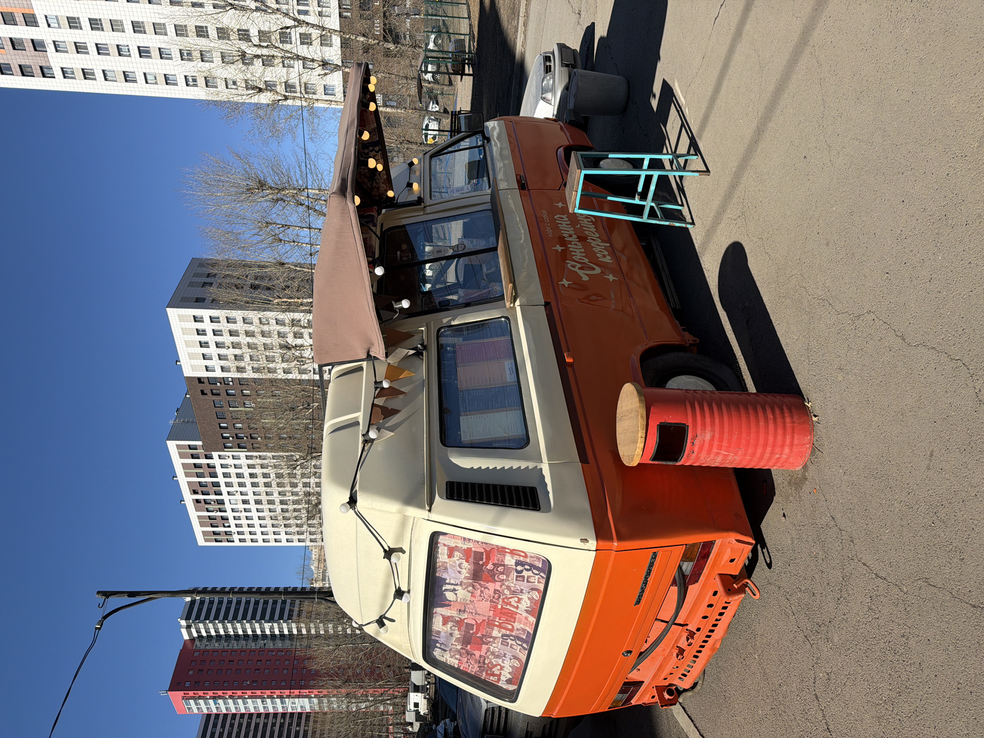 The image shows a retro-style orange and white van, seemingly a mobile coffee shop, parked on a paved surface in what appears to be Krasnoyarsk, Russia. Buildings with many windows are visible in the background, suggesting an urban setting. The van has a brown awning with decorative lights. A red barrel is used as a makeshift table next to the van, and a teal-colored metal structure also serves as a table. The van has writing in Russian on its side.