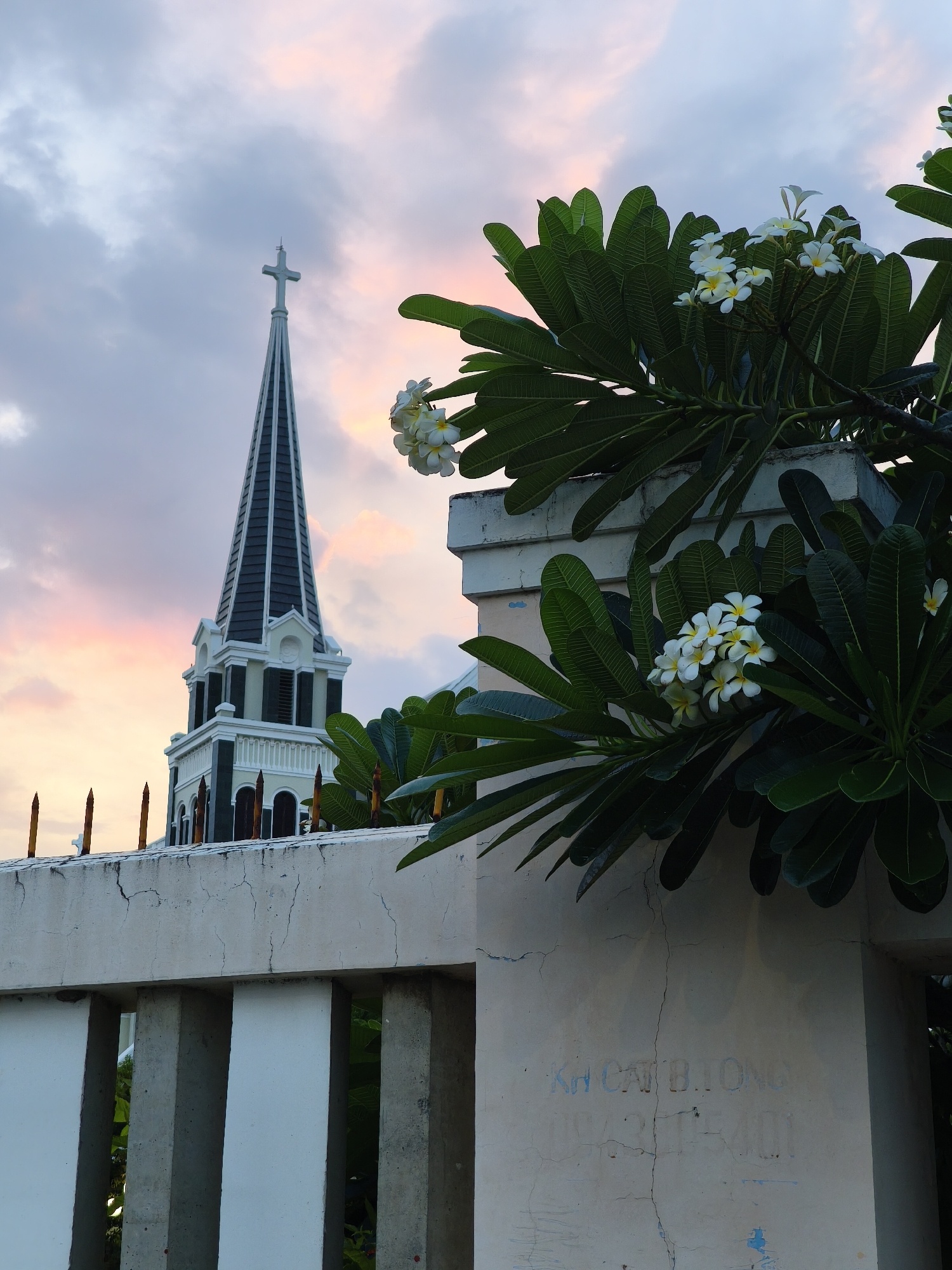 The image shows a church steeple in Bac Nha Trang Ward, Vietnam, under a twilight sky with soft pink and grey clouds. In the foreground, a white concrete wall with vertical slits and a textured surface is visible. On the right side of the wall, there is some faint blue lettering and a vertical crack. A branch of a tree with large green leaves and white flowers (likely frangipani) extends into the frame from the right, partially obscuring the church. The church steeple is tall and slender, with a dark roof and a white cross at its apex. The steeple has multiple levels with arched windows and decorative white trim. The overall scene suggests a serene and perhaps religious setting in Vietnam.