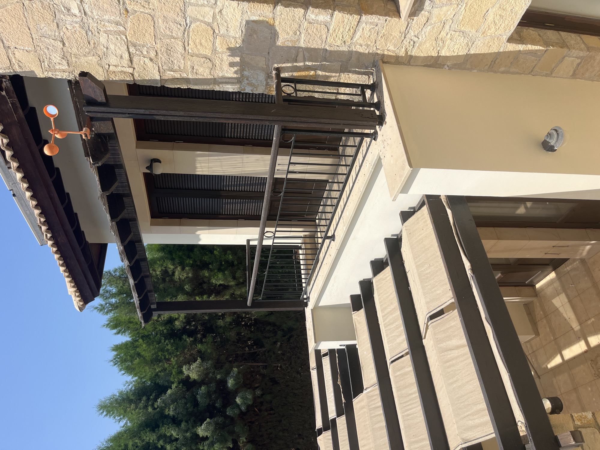 The image shows an exterior view of a building with a tiled roof and stone walls, located in Aphrodite Hills, Cyprus. In the foreground, there is a covered patio area with dark wooden beams and beige fabric awnings. Above this, a balcony with a dark metal railing overlooks the patio. The balcony is accessed through a door with dark shutters. A small orange weather vane is mounted on the wooden structure above the balcony. The background features a dense line of green cypress trees against a clear blue sky. The overall scene depicts a sunny day in a Mediterranean architectural setting.
