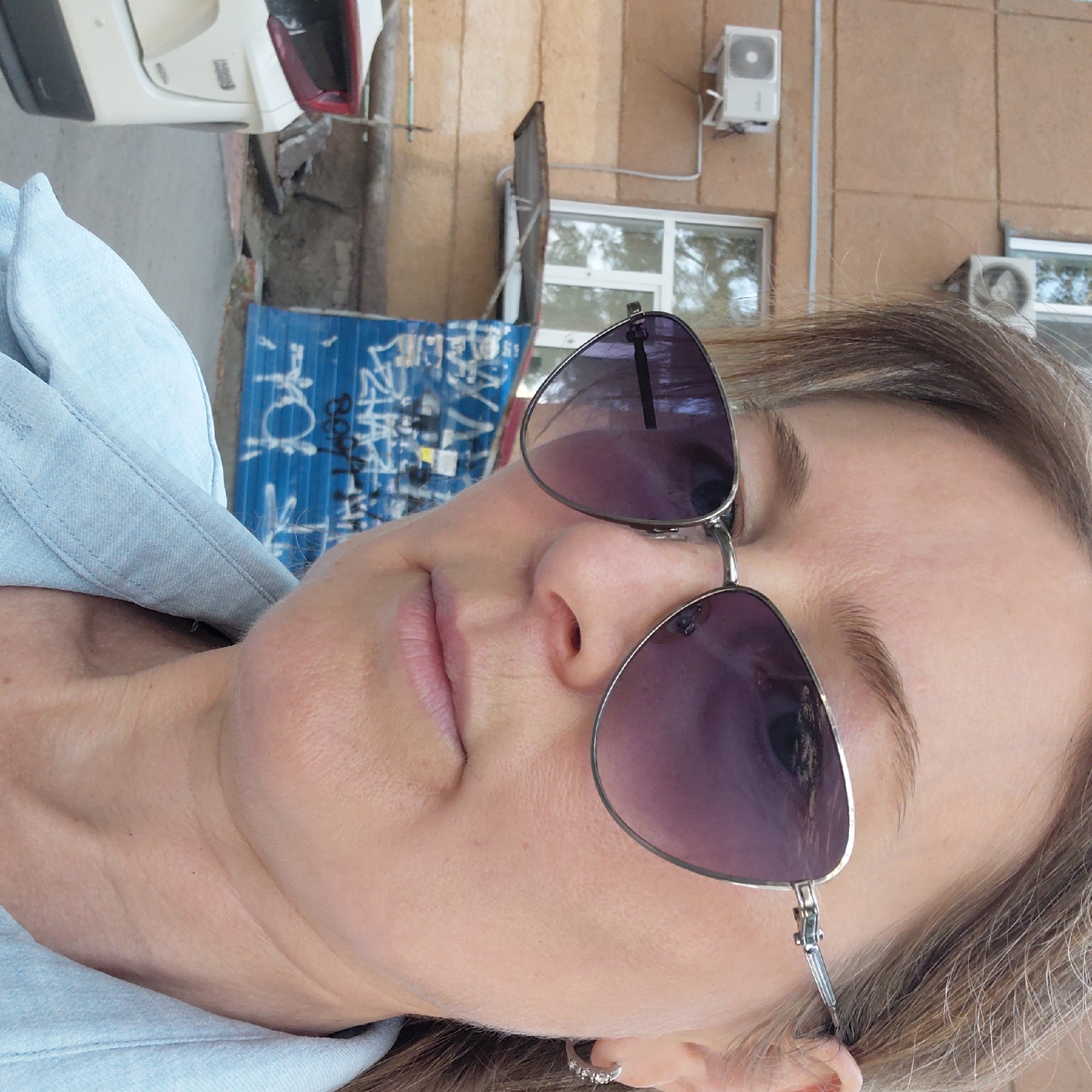 A woman with light brown hair and sunglasses is taking a selfie in front of a building. She is wearing a light blue denim shirt. The building behind her is light brown with white window frames and air conditioning units. There is graffiti on a blue wall in the background, and a white car is partially visible on the left side of the frame. This scene appears to be in an urban environment in Novosibirsk, Russia.